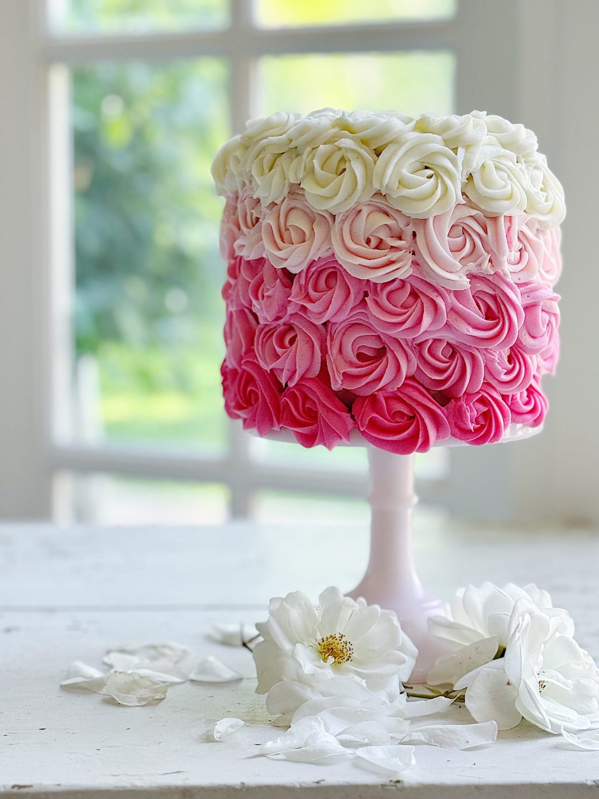 A tall cake decorated with ombre pink and white buttercream rosettes sits on a white stand, with white flowers and petals on the table in front.