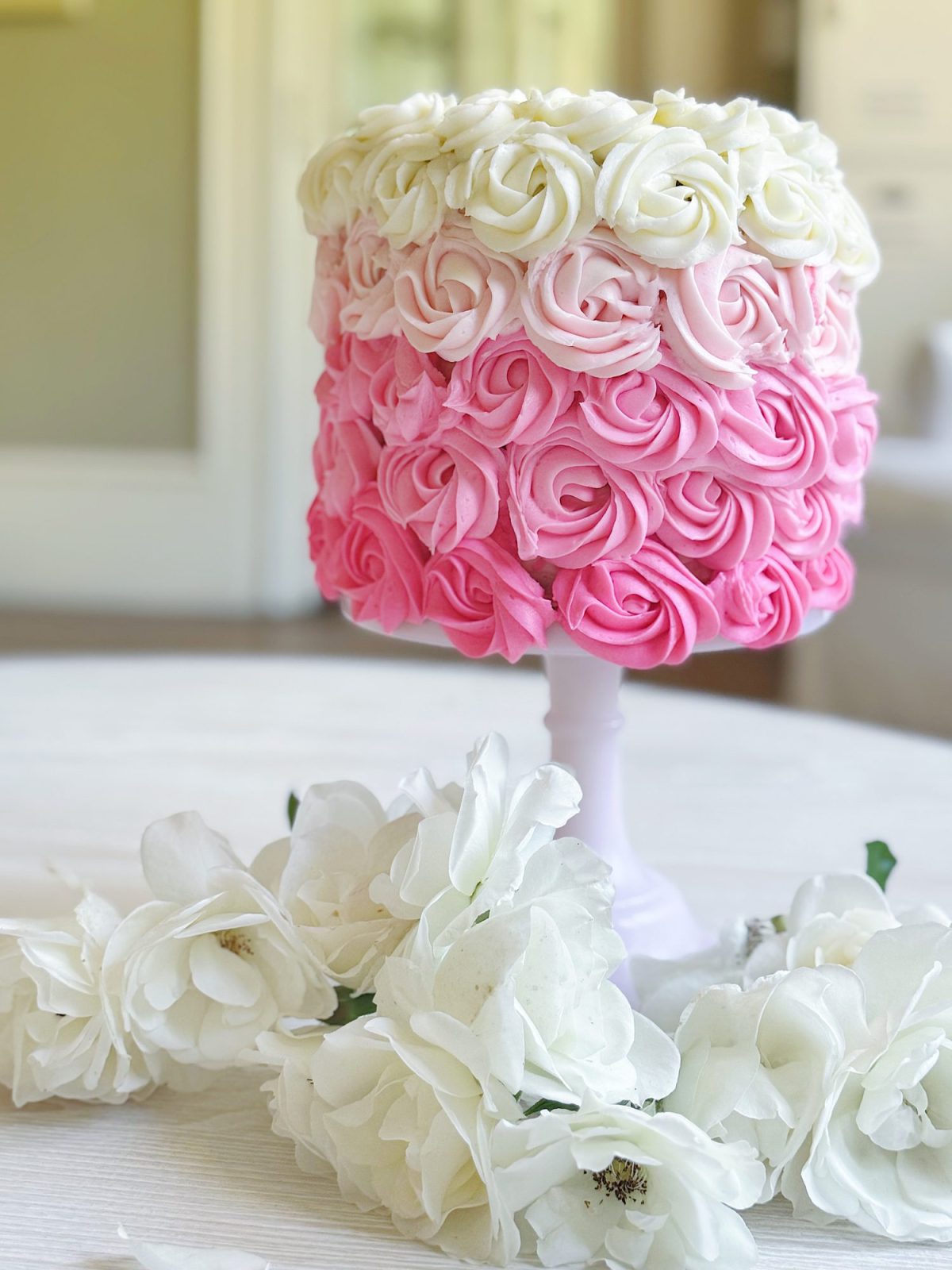 A cake decorated with white and pink rosette icing sits on a white cake stand, surrounded by white artificial flowers on a table.