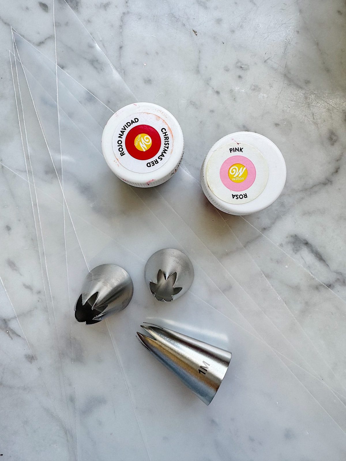 Two containers of food coloring, three metal piping tips, and several clear piping bags are arranged on a marble surface.