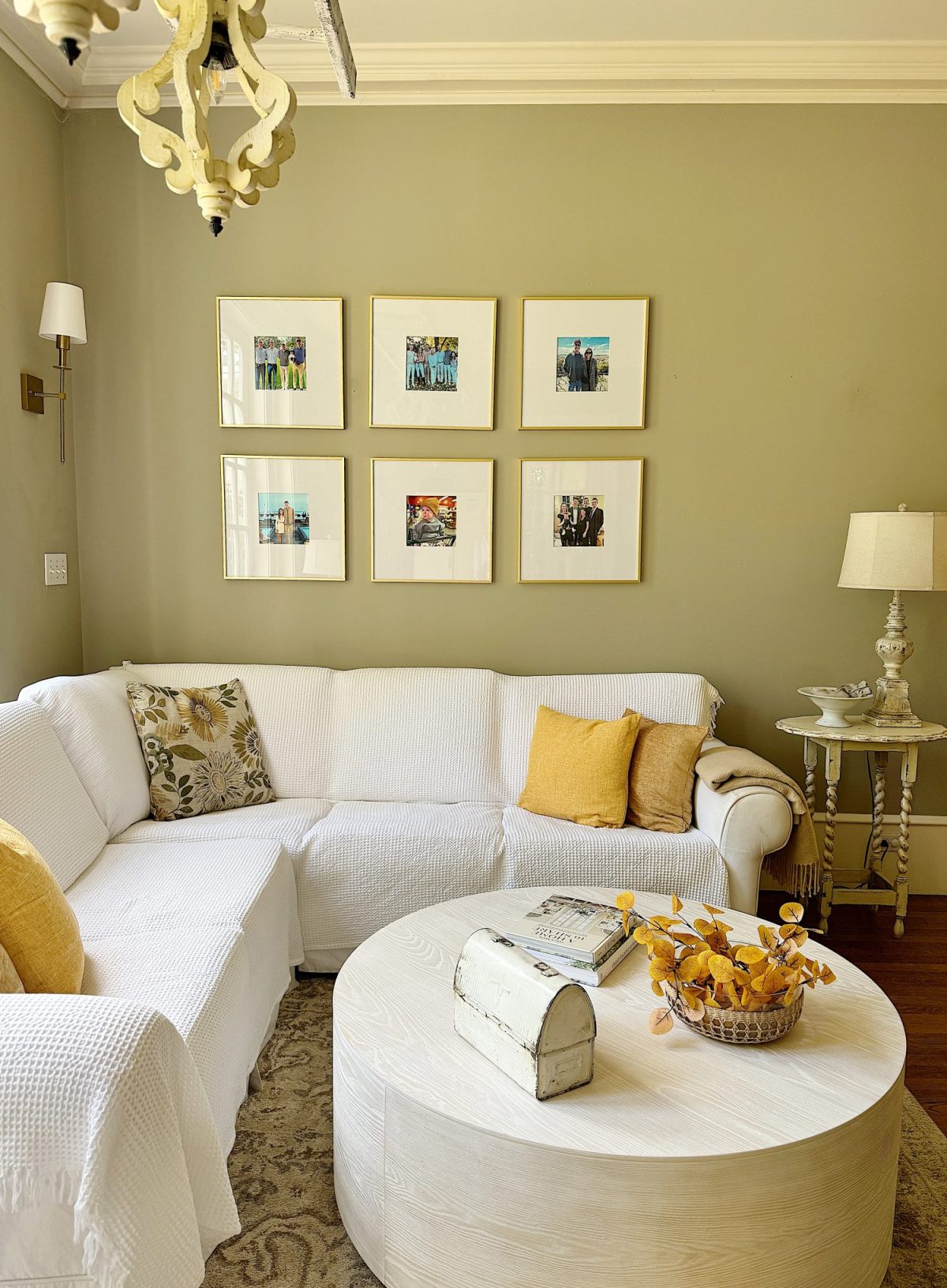 A living room with a white sectional sofa, yellow and floral pillows, a round white coffee table, wall art, a lamp on a side table, and a decorative chandelier.