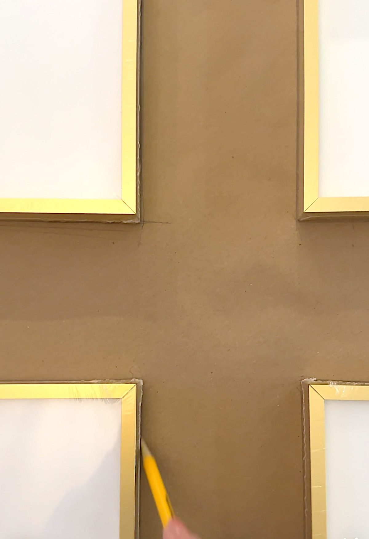 Four gold-framed pictures are arranged in a grid on a brown surface. A hand holding a pencil traces the corner of one frame.