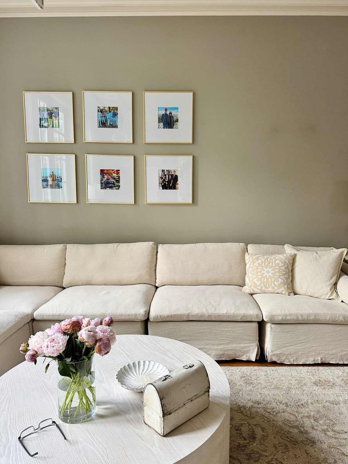 A beige sectional sofa with patterned pillows sits against a taupe wall displaying six framed photos. A round coffee table holds a vase of pink flowers, a decorative box, and a shell-shaped dish.