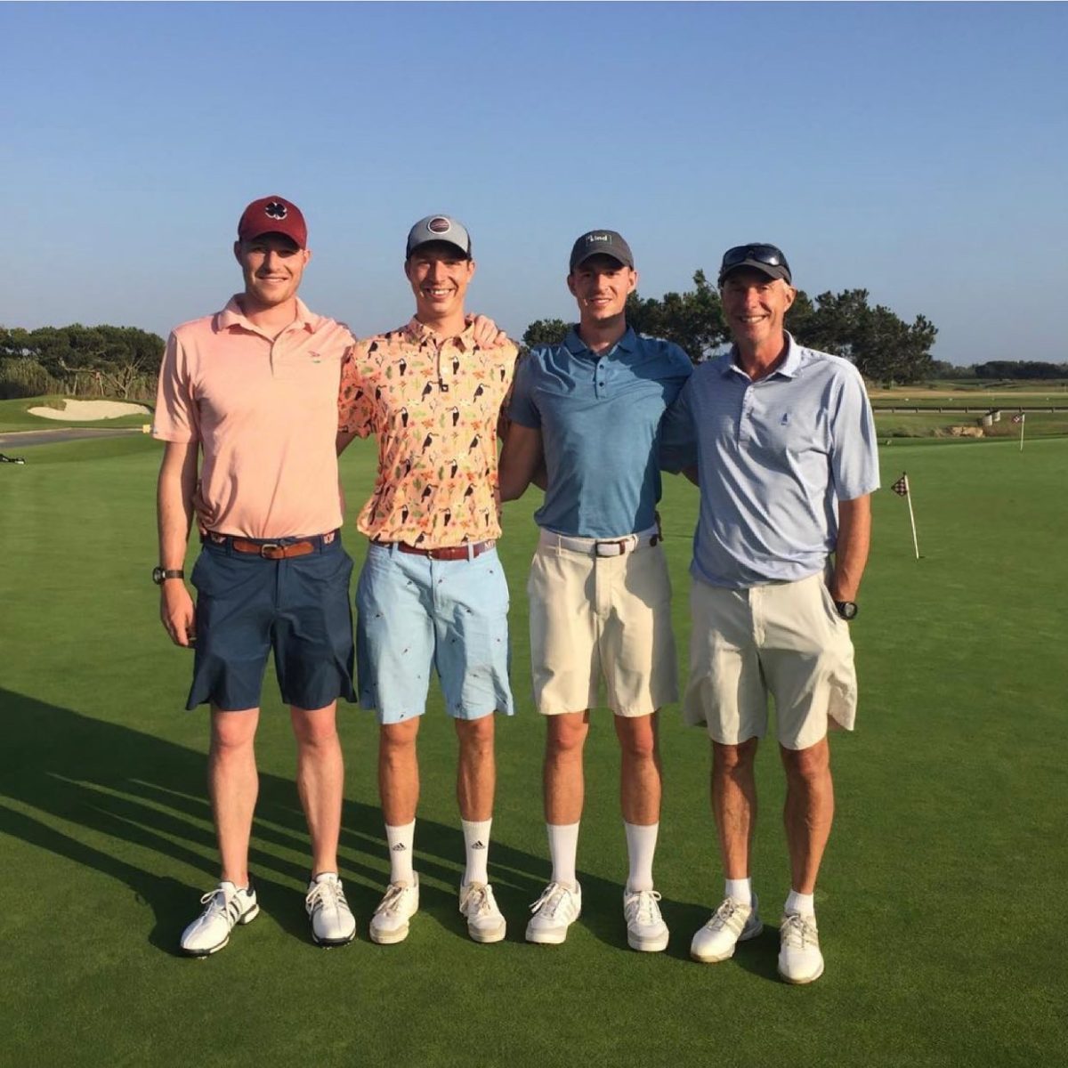 Four men standing side by side on a golf course green, wearing golf attire and caps, smiling at the camera.