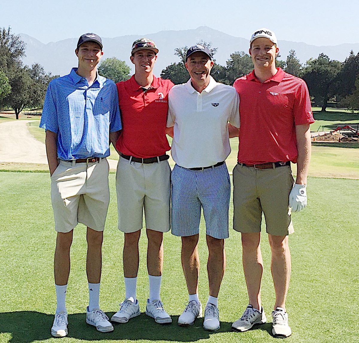 Four men stand side by side on a golf course, dressed in golf attire with hats and shorts, posing for a group photo with a scenic mountain backdrop.
