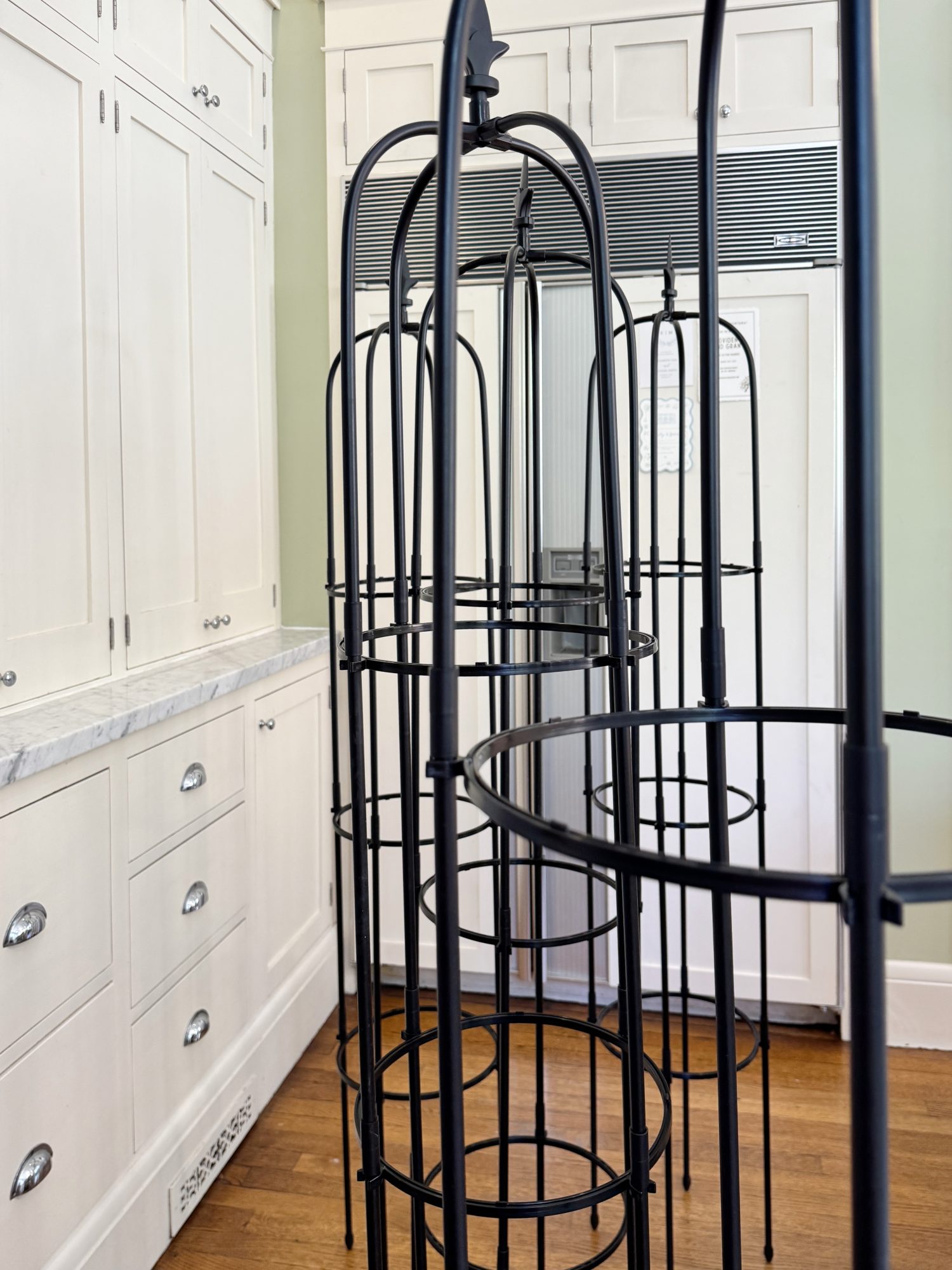 Three black metal garden trellises stand upright indoors on a wooden floor, next to white kitchen cabinets and drawers.