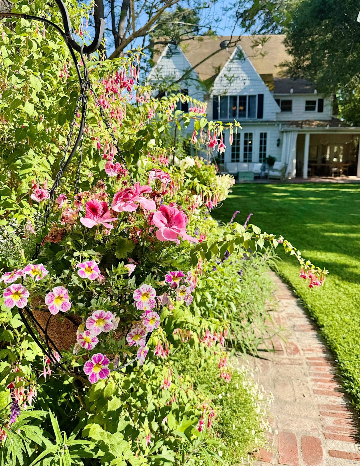 Pink and purple flowers line a brick pathway leading to a white house with steep gables and large windows, surrounded by green lawn and trees on a sunny day.