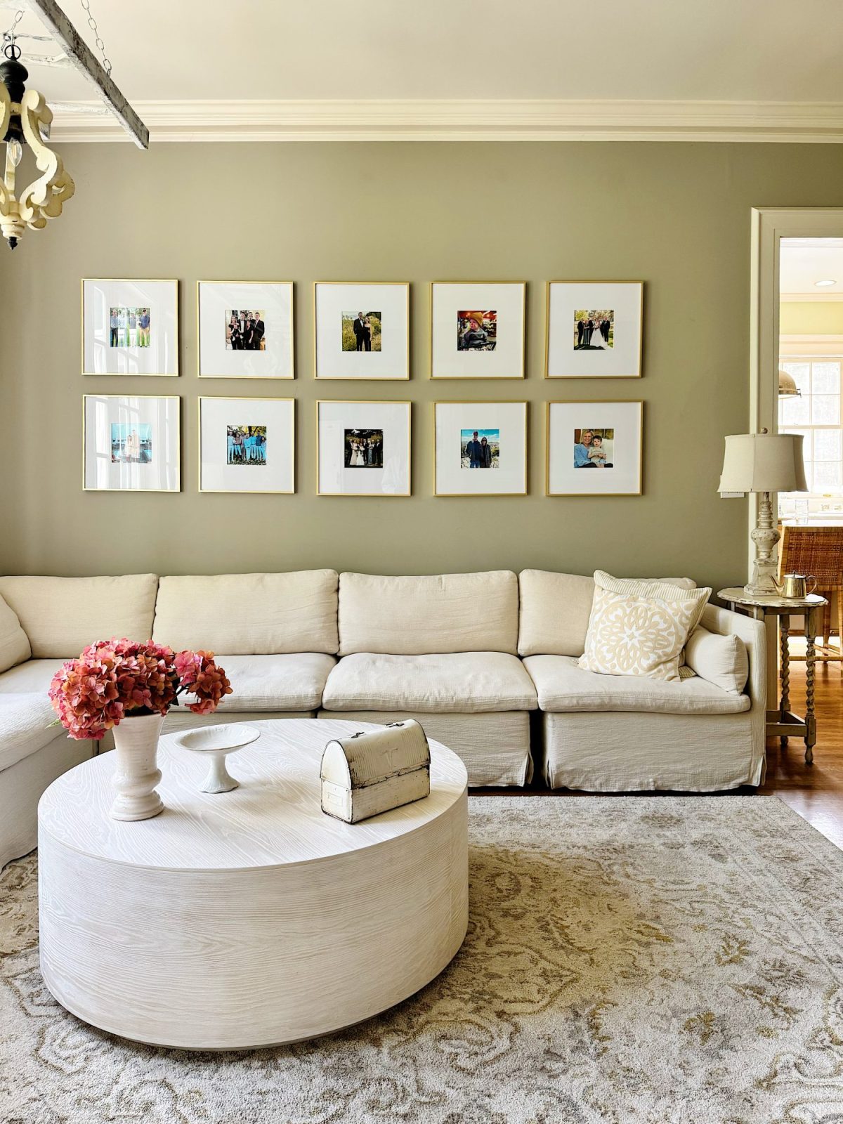 A living room with a white sectional sofa, round white coffee table, flower arrangement, and framed photos arranged in two rows on a beige wall.