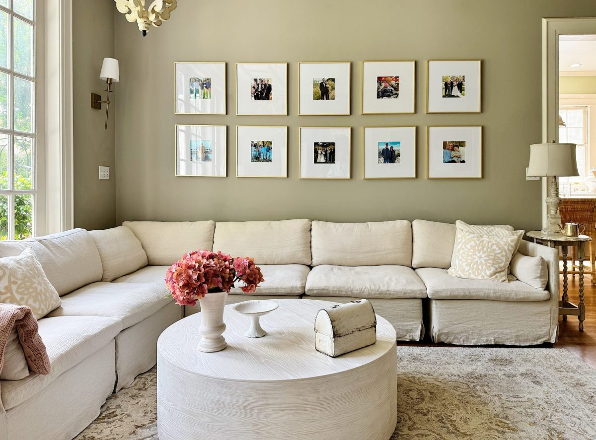 A neutral-toned living room with a large white sectional sofa, a round white coffee table, and framed photos arranged in two rows on the beige wall.