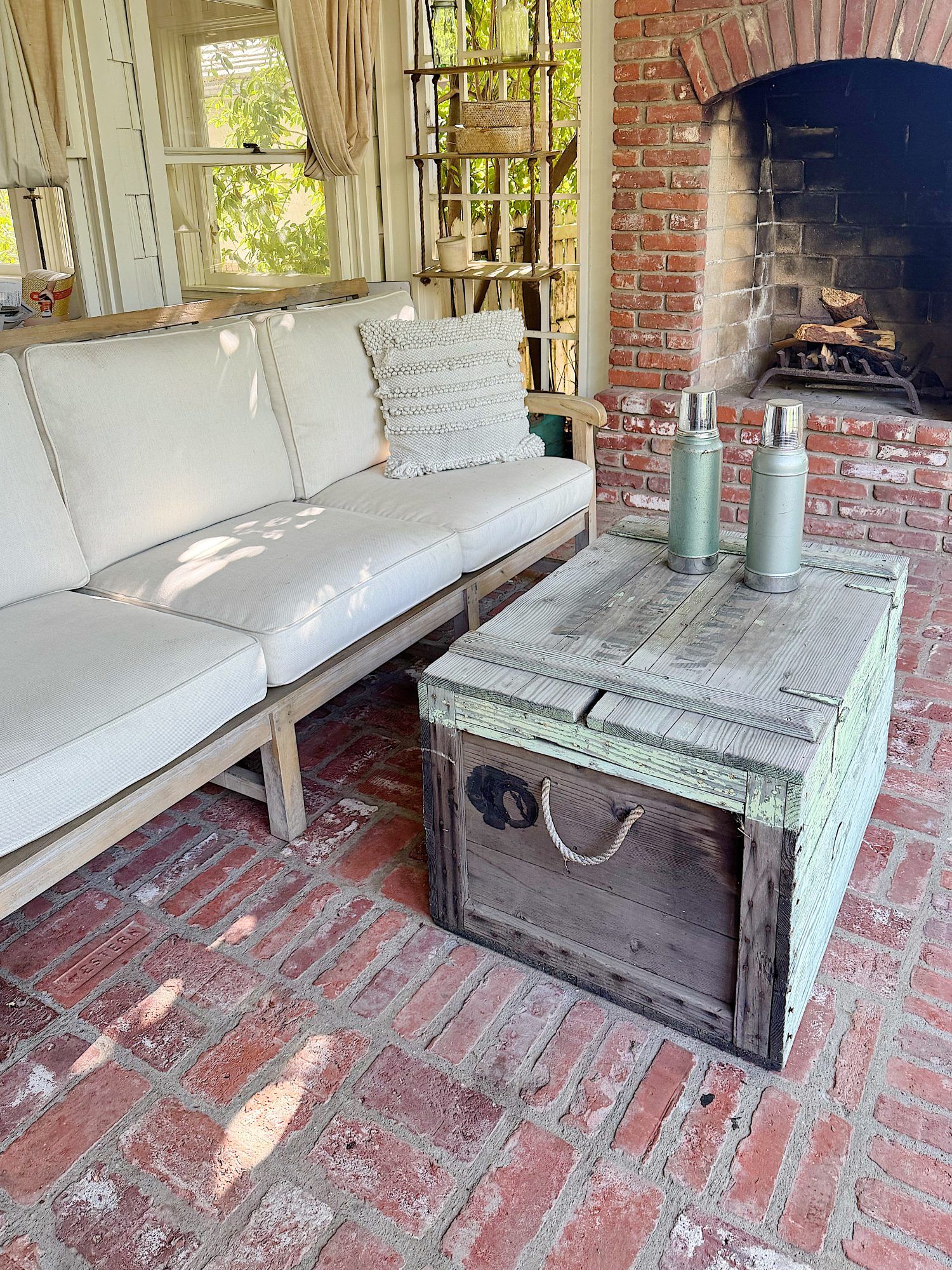 A beige cushioned sofa sits next to a rustic wooden trunk coffee table with two metal bottles on top, on a brick floor near a brick fireplace.