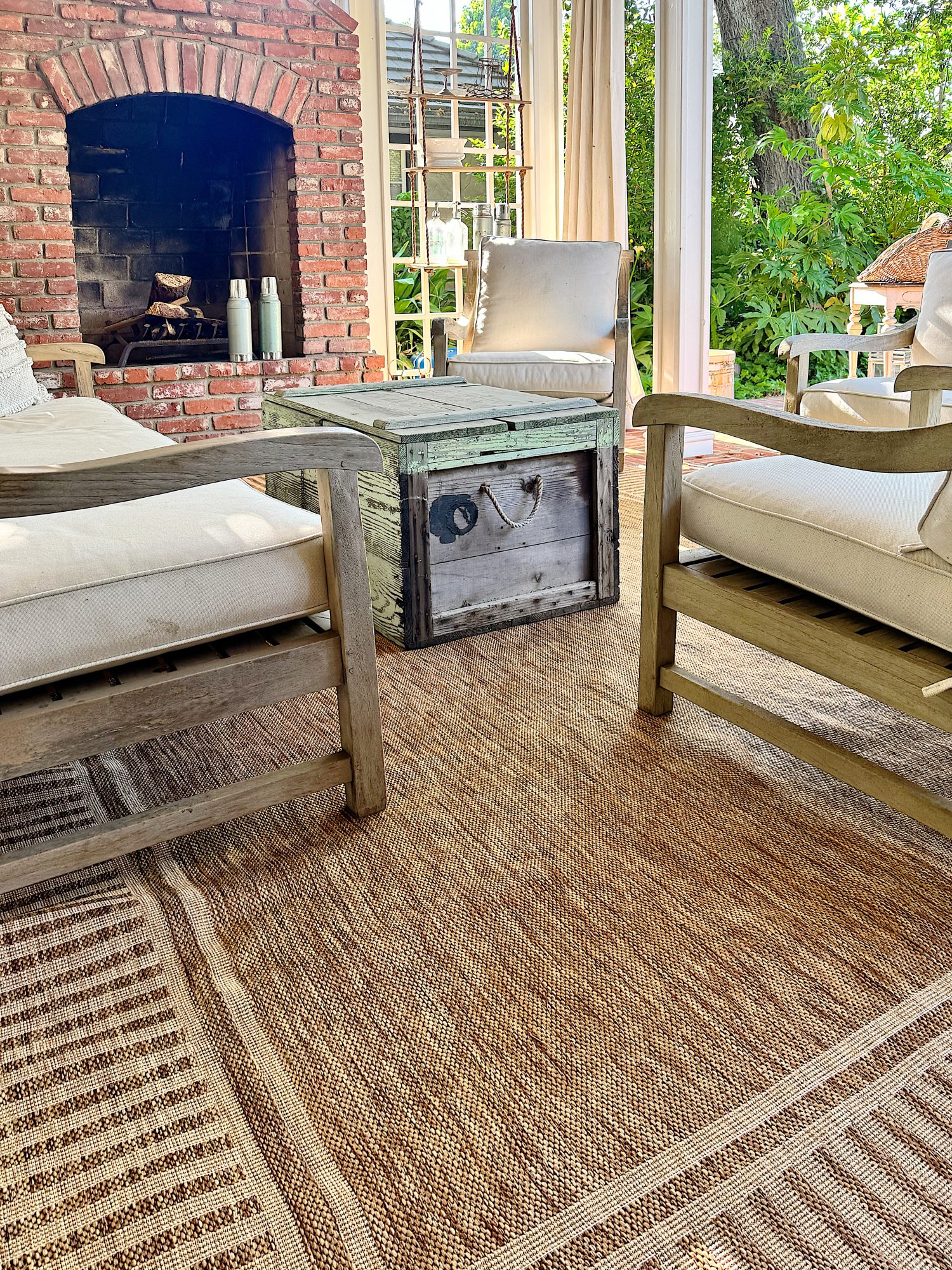 Outdoor patio with cushioned wooden chairs, a wooden coffee table, and a brick fireplace. The area is covered with a woven rug and surrounded by greenery.