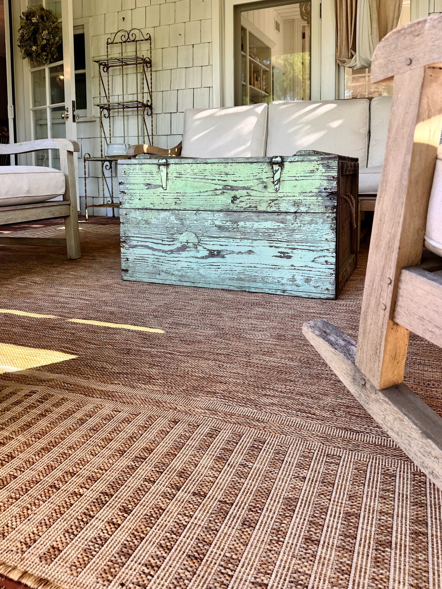 A weathered green wooden chest is used as a coffee table on a covered porch with wicker furniture and a brown woven rug.