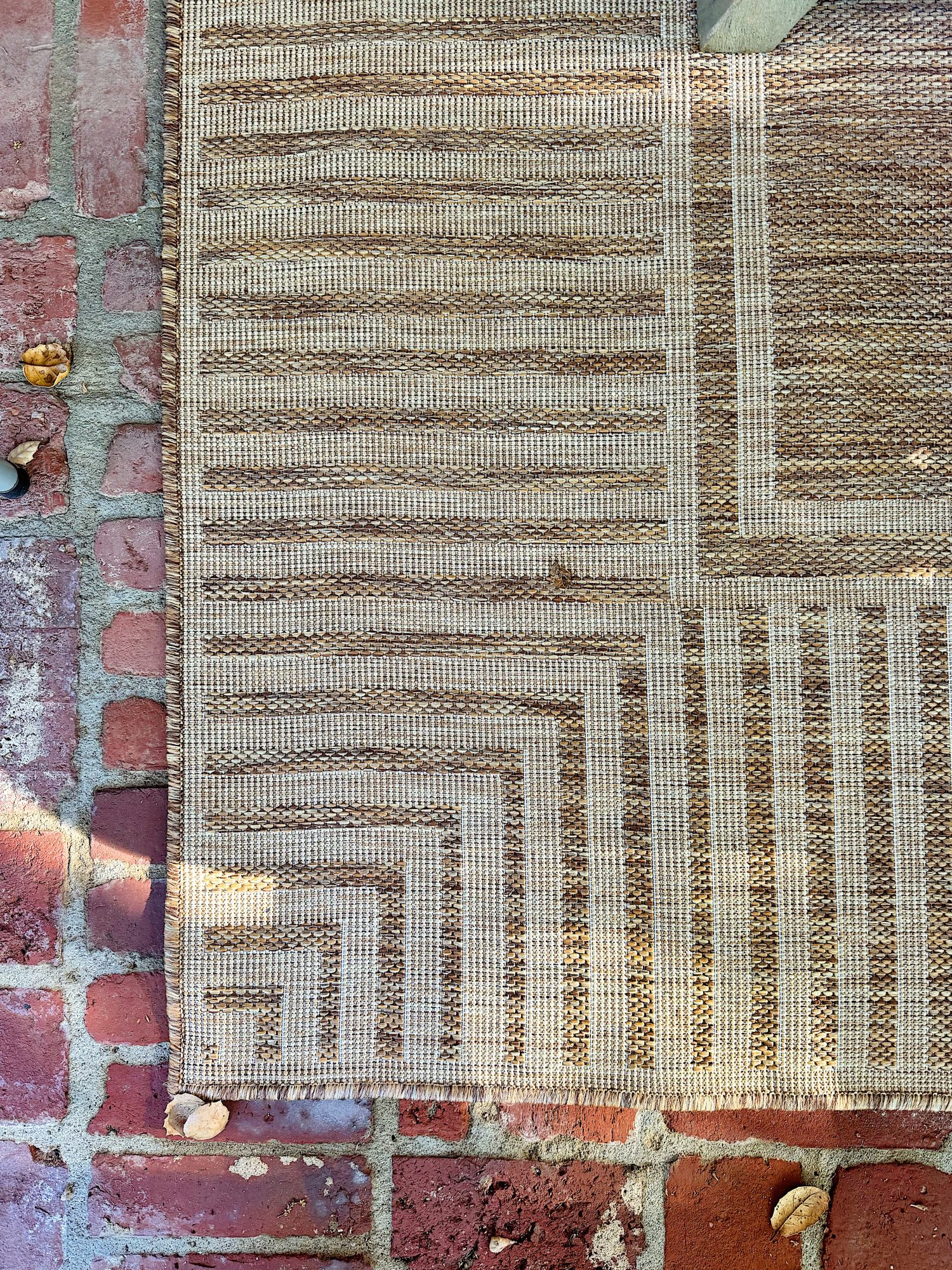 A beige patterned rug with geometric lines lies on a red brick surface. A few small leaves are scattered on the rug and bricks.