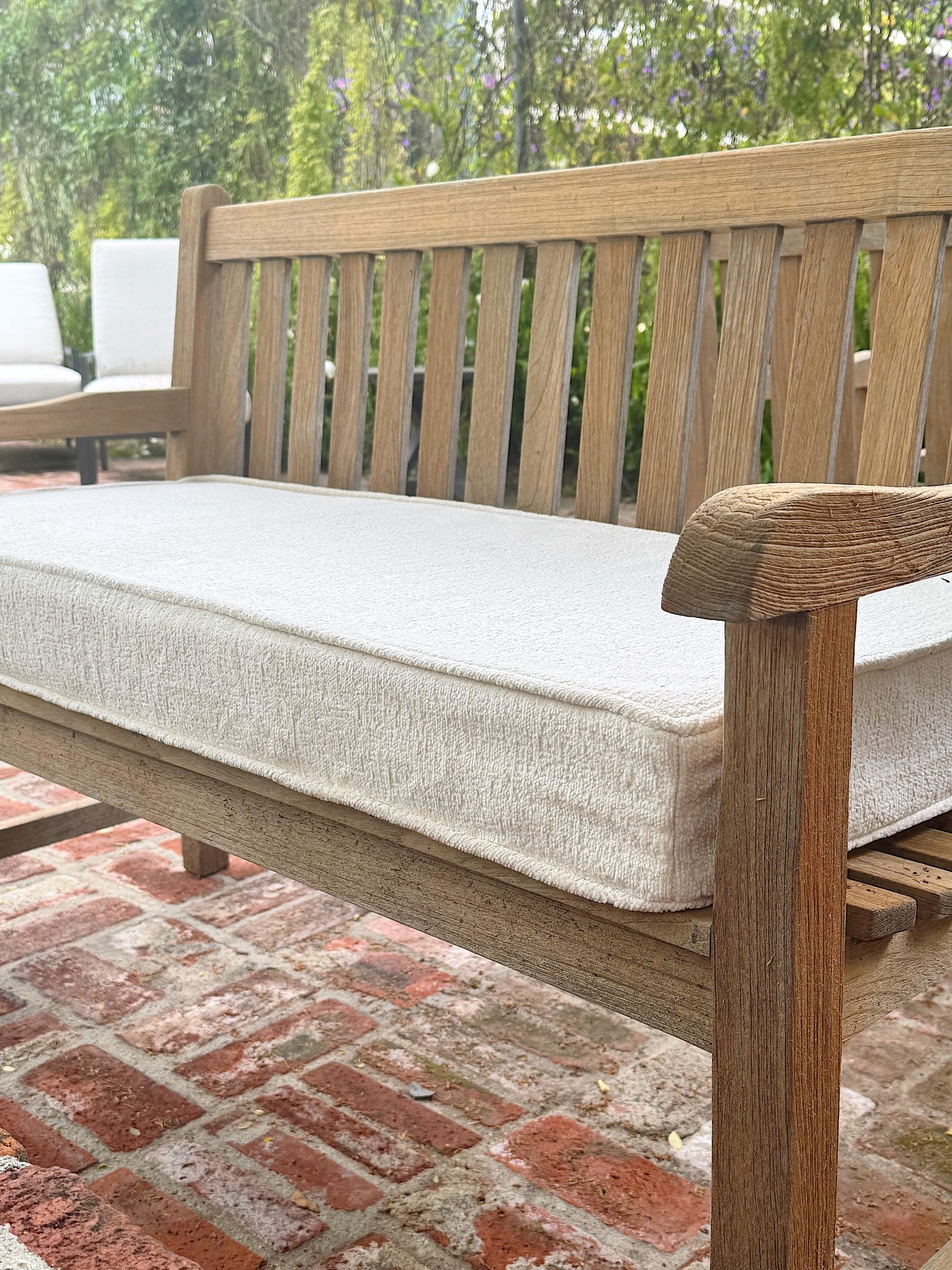 Wooden outdoor bench with a thick light-colored cushion on a red brick patio, with greenery and additional seating in the background.