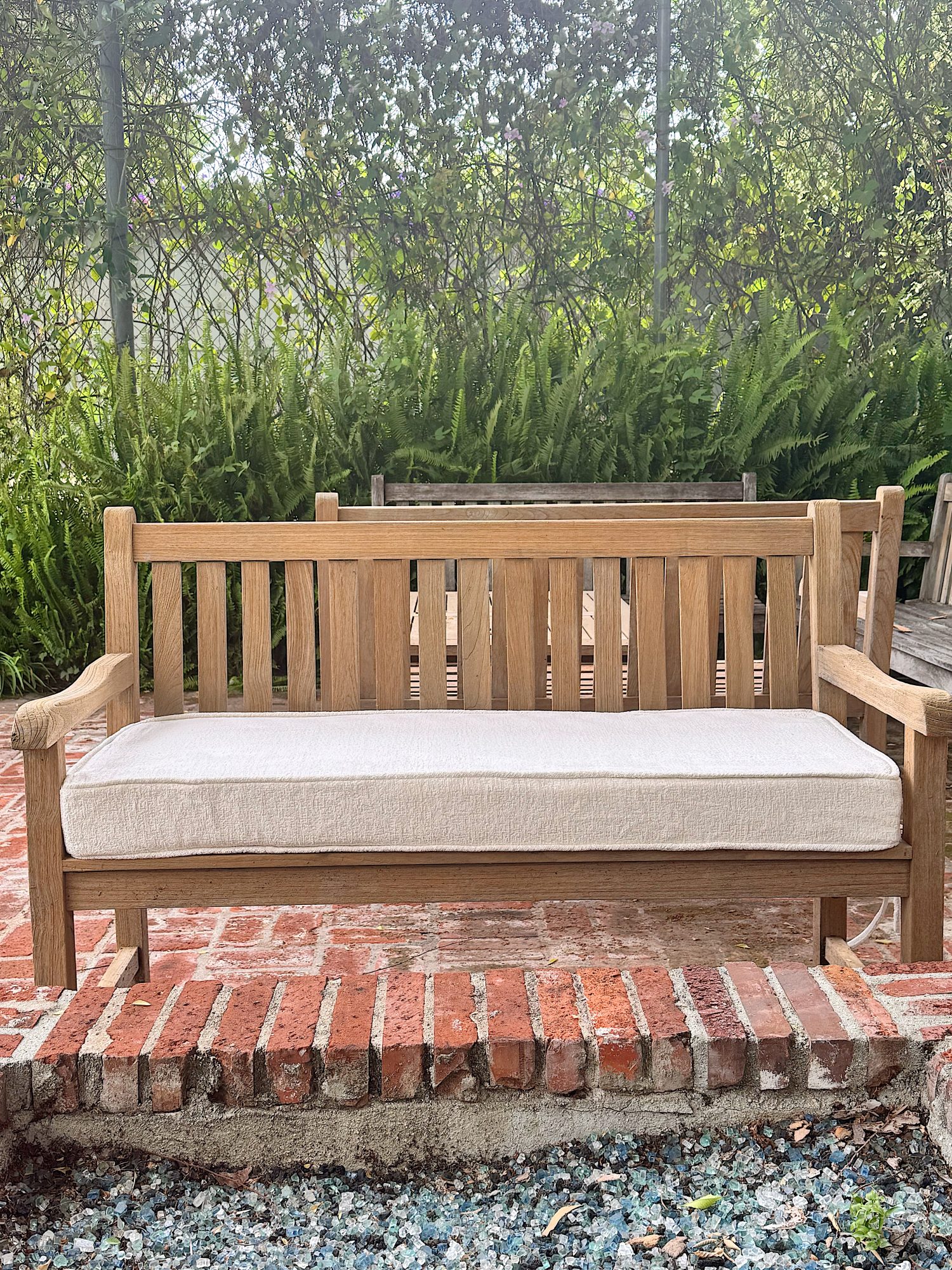 A wooden outdoor bench with a beige cushion sits on a brick patio, surrounded by greenery and ferns in the background.
