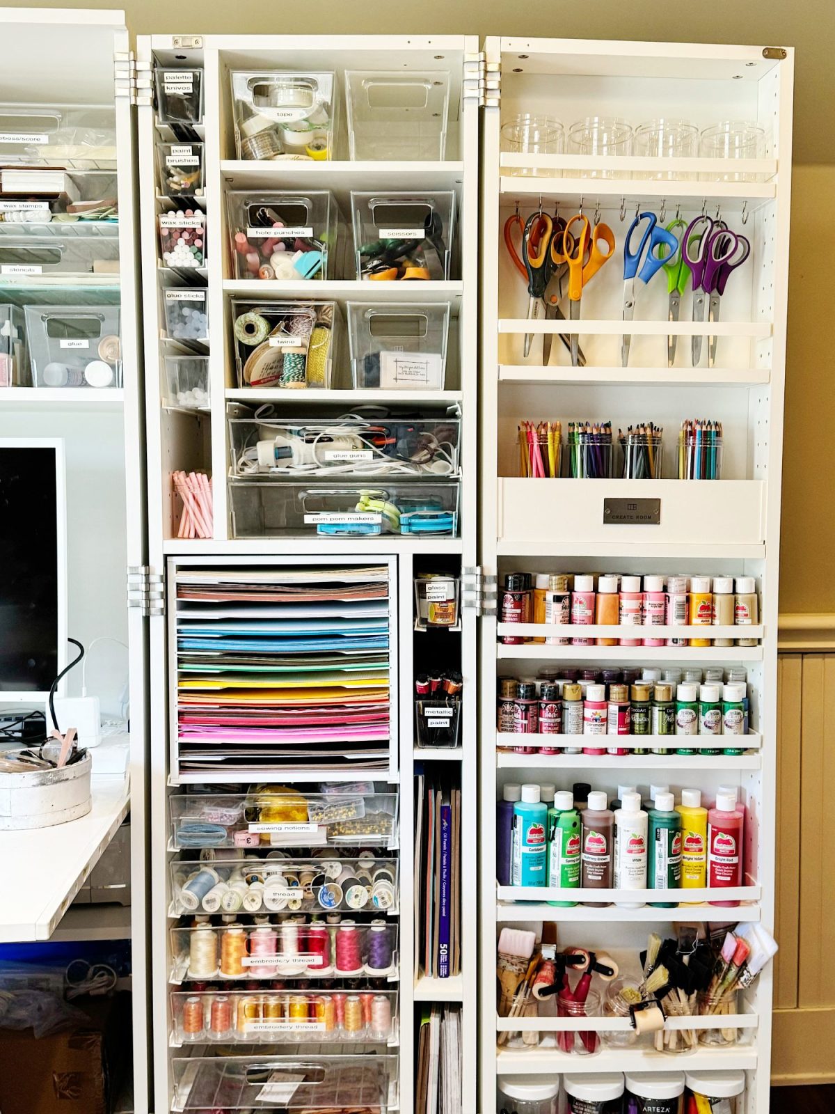 Organized white shelving unit with various craft supplies, including paper, paint bottles, scissors, ribbons, thread, and storage bins, neatly arranged by category.