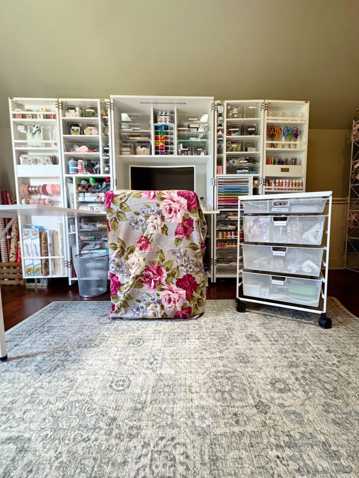 A craft room features organized white storage cabinets filled with supplies, a floral-covered chair, a rolling drawer cart, and a light-colored patterned rug.