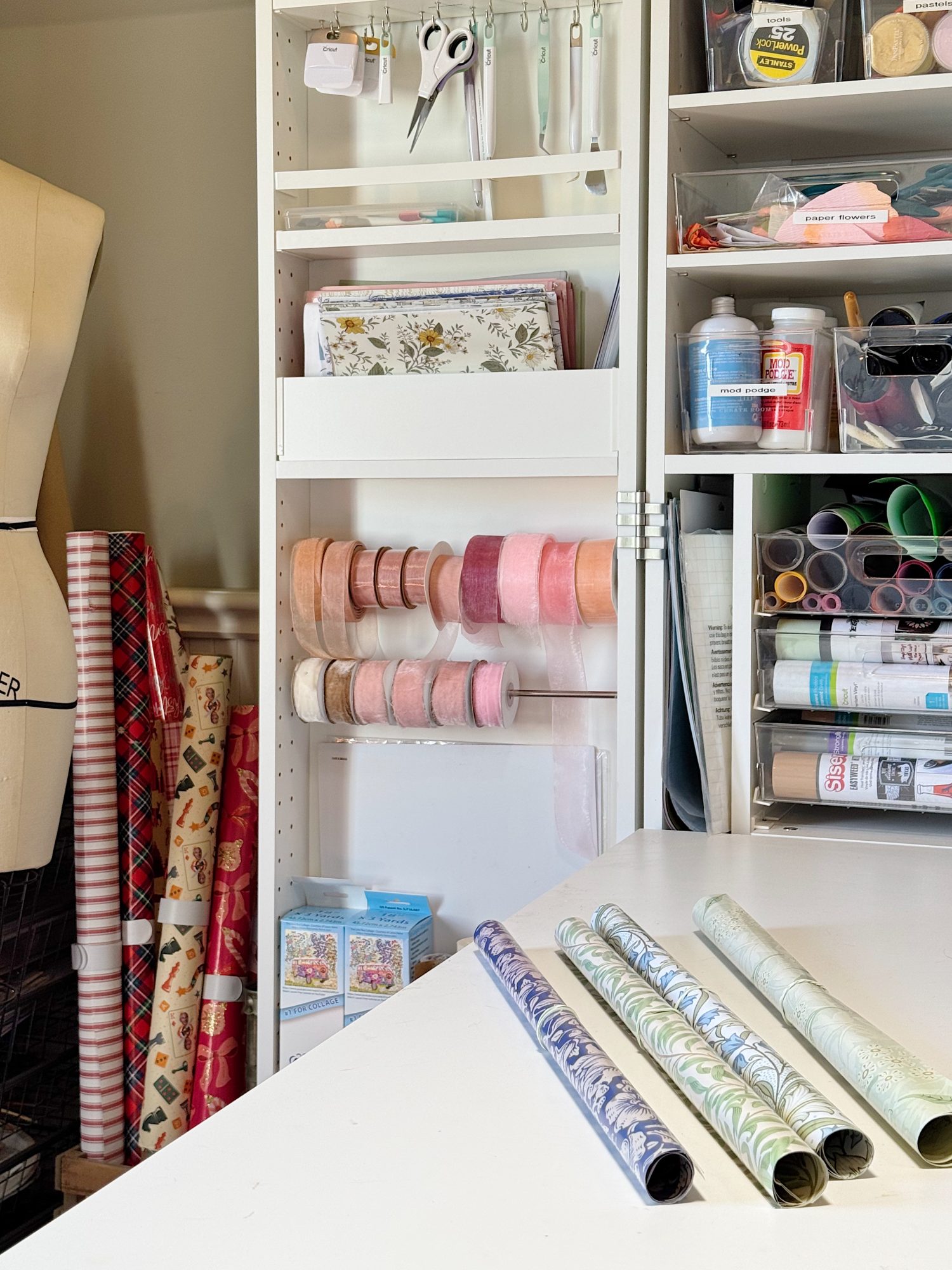 A craft room with rolls of wrapping paper, ribbons, scissors, paint, fabric, and other art supplies neatly organized on shelves and a table.
