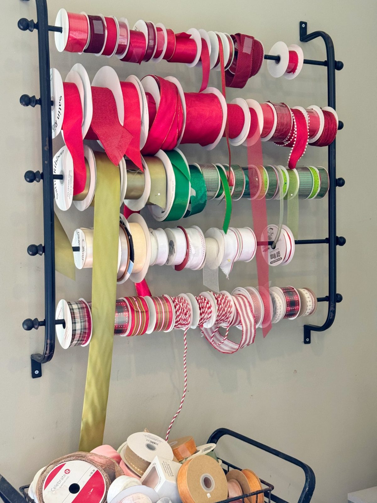 A wall-mounted rack holds multiple spools of ribbon in various colors and patterns, with additional spools of ribbon arranged on a cart below.