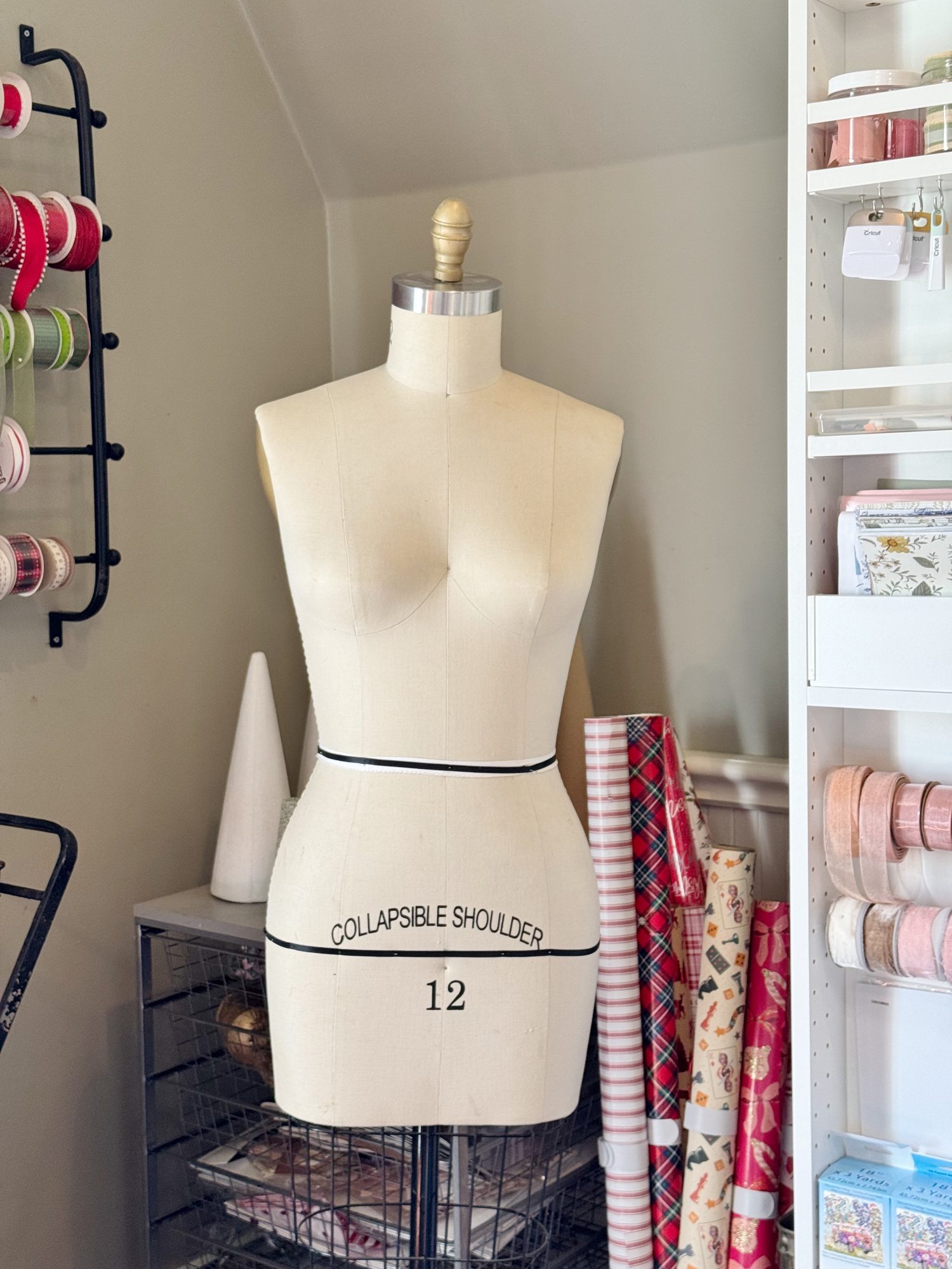 A dress form labeled "Collapsible Shoulder 12" stands in a craft room beside shelves of ribbon and rolls of wrapping paper.