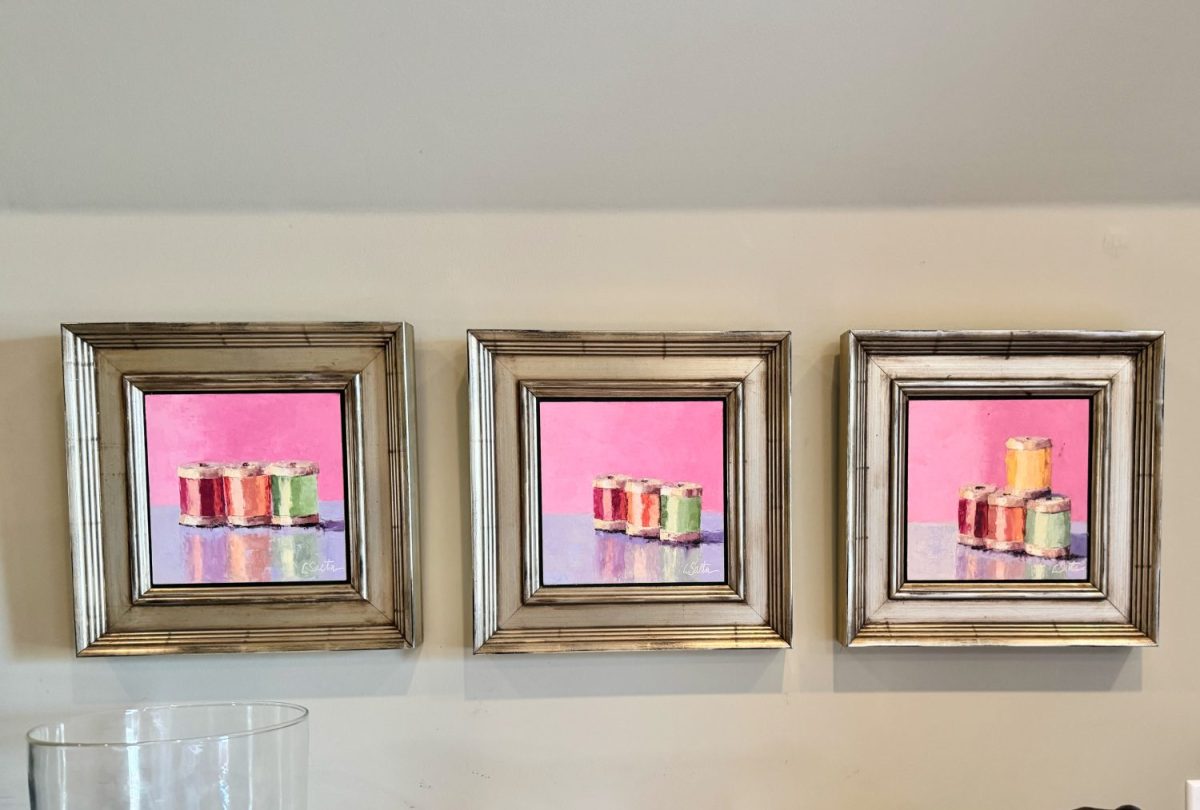 Three framed paintings of colorful stacked cylinders on reflective surfaces, each against a bright pink background, are displayed side by side on a light-colored wall.