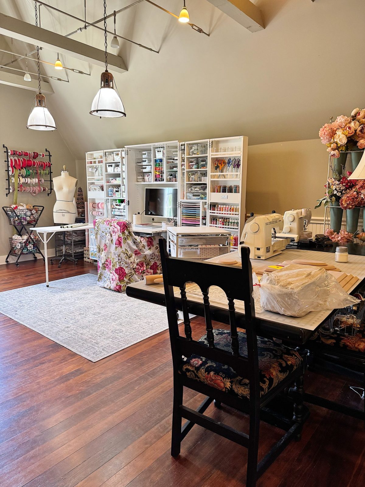 A well-organized sewing room with shelves of supplies, a sewing machine, dress form, floral decor, and wooden floors, viewed from a table with a black chair in the foreground.