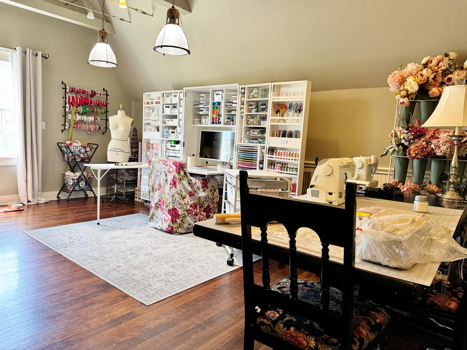 A well-organized craft room with sewing machines, shelves of supplies, a dress form, and floral decor on hardwood floors under pendant lights.