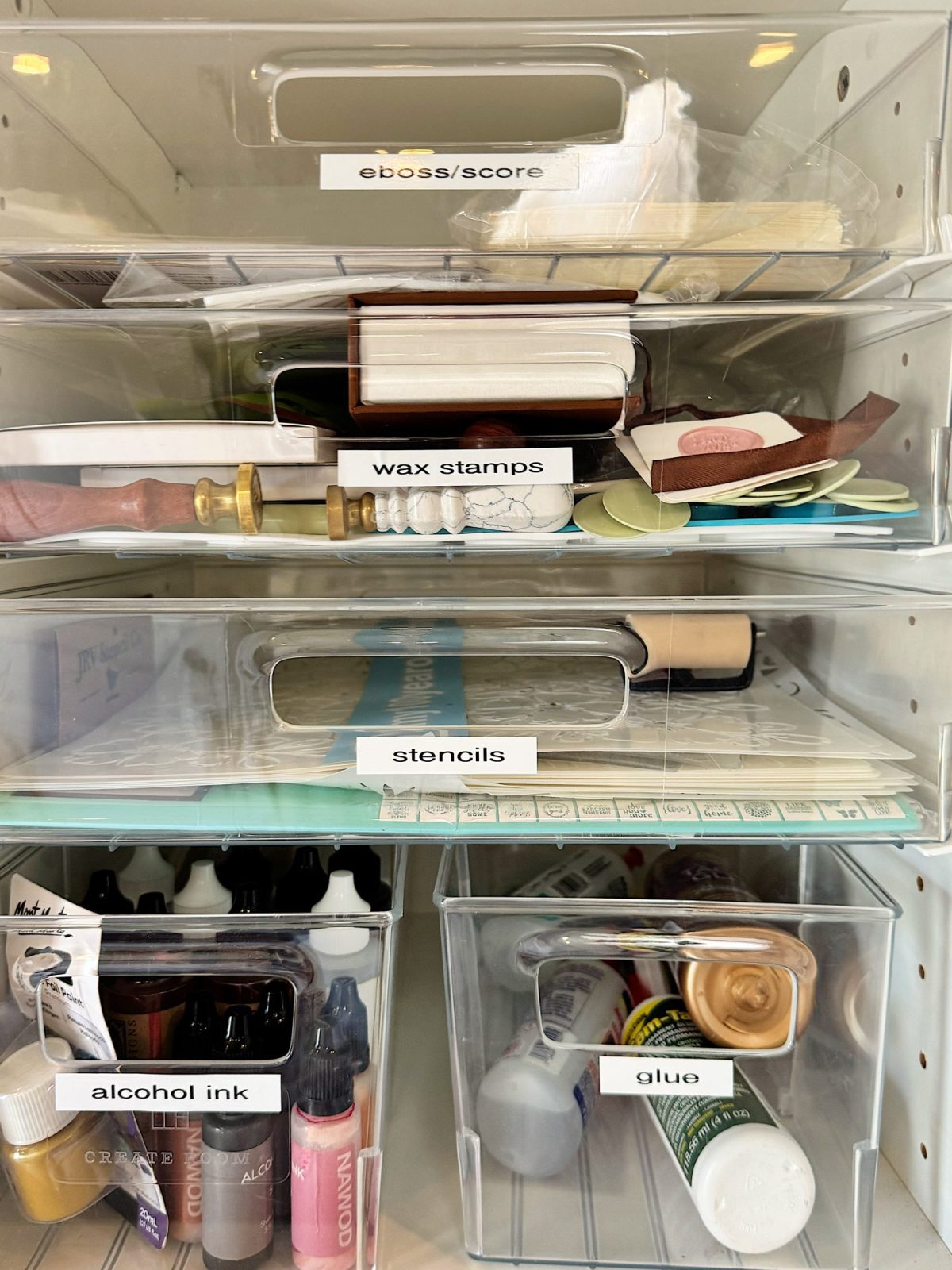 Clear storage bins labeled "emboss/score," "wax stamps," "stencils," "alcohol ink," and "glue," each containing various crafting supplies, organized on shelves.