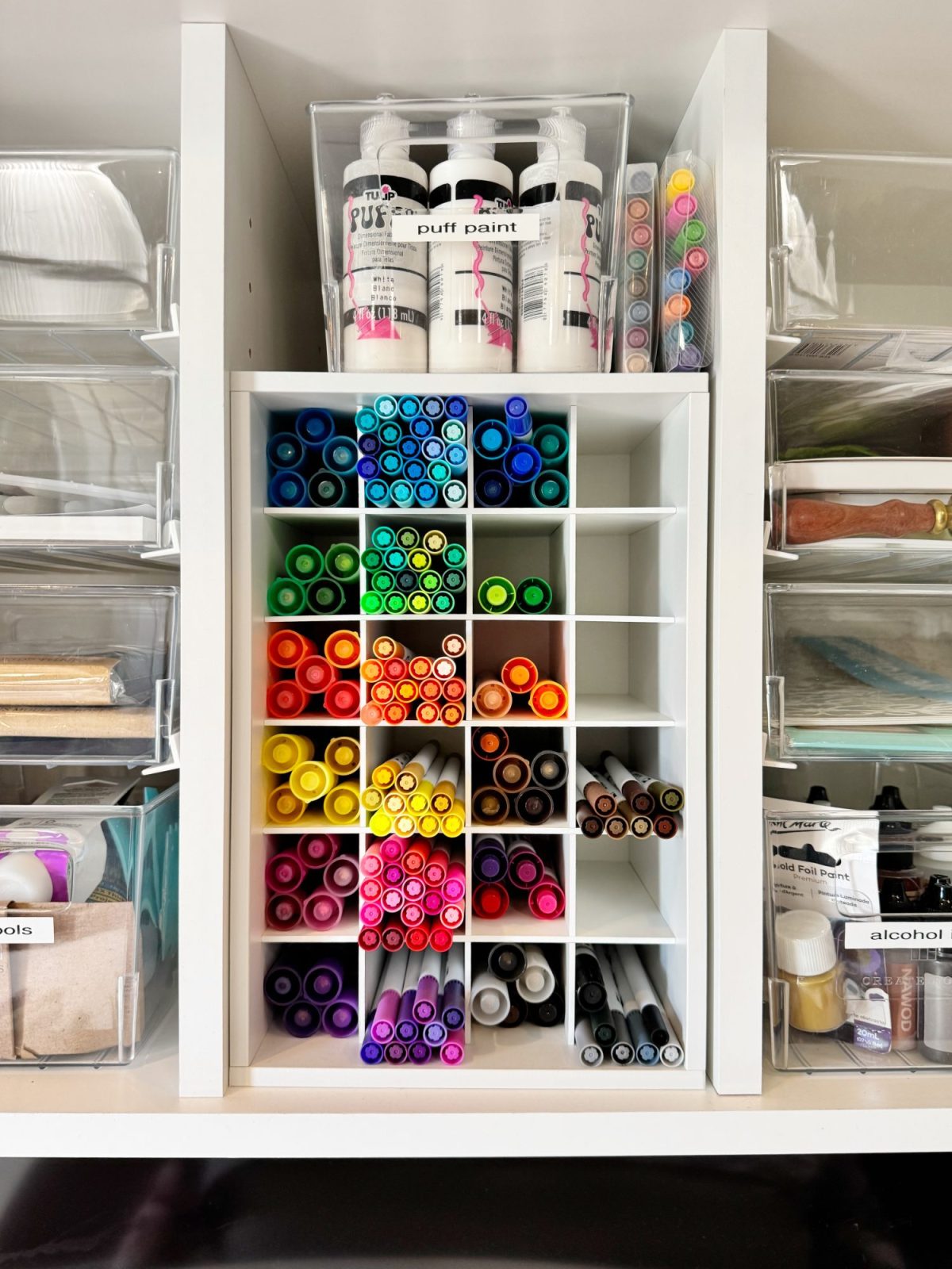 A white cubby organizer holds markers arranged by color, with bottles of paint and containers of art supplies on shelves above and around it.