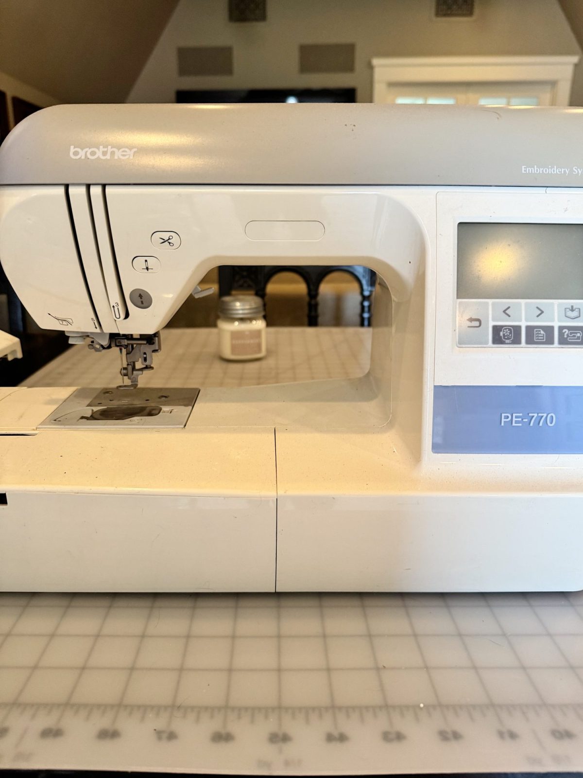 A Brother PE-770 embroidery sewing machine sits on a grid-patterned table, with a candle and chairs in the background.