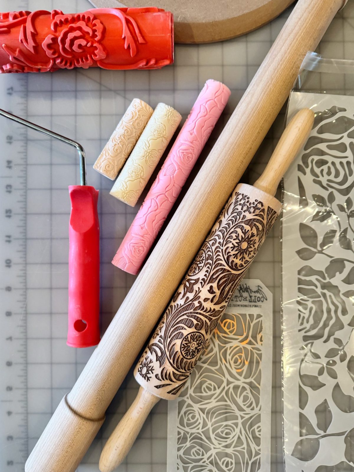 Various rolling pins, texture rollers, embossing mats, and stencils with floral and decorative patterns are arranged on a gridded cutting mat.