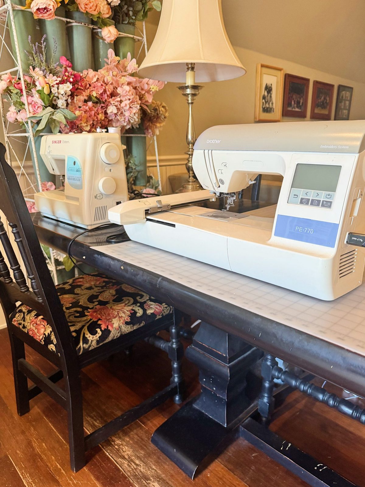 Two sewing machines sit on a table with a floral arrangement, a chair, and a lamp in a well-lit room with framed pictures on the wall.