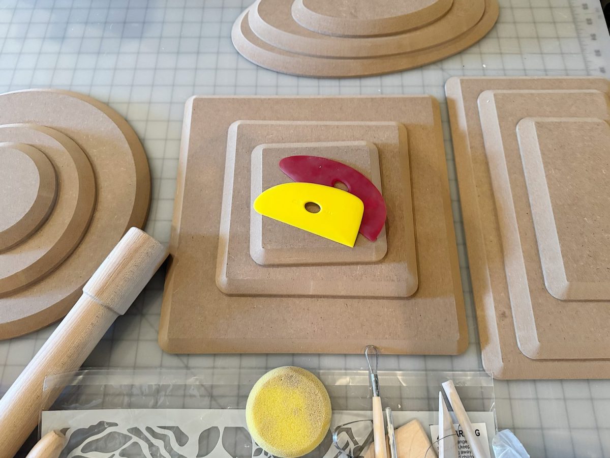 Assorted pottery tools and wooden templates arranged on a gridded work surface, including a rolling pin, sponge, and two plastic scrapers, one yellow and one red.