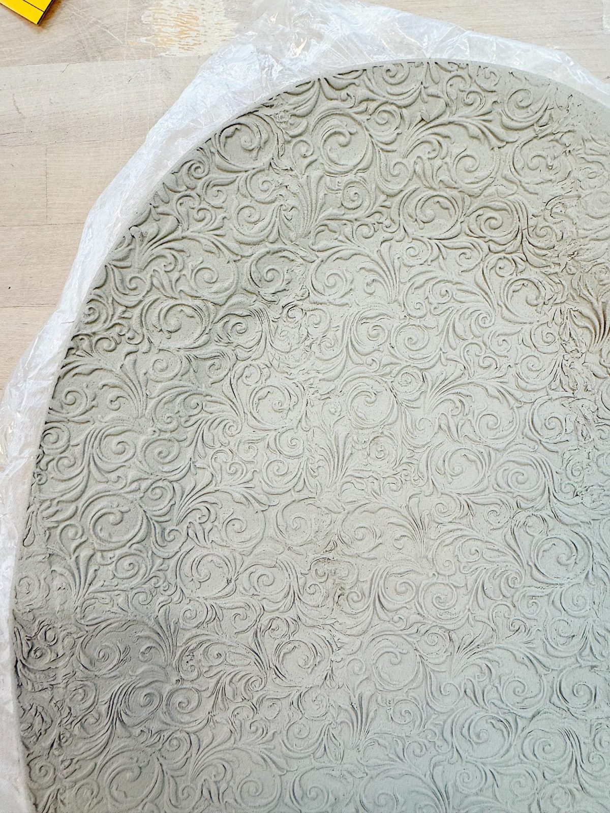 A round slab of clay with an intricate, raised swirling pattern on its surface, partially covered with a sheet of clear plastic.