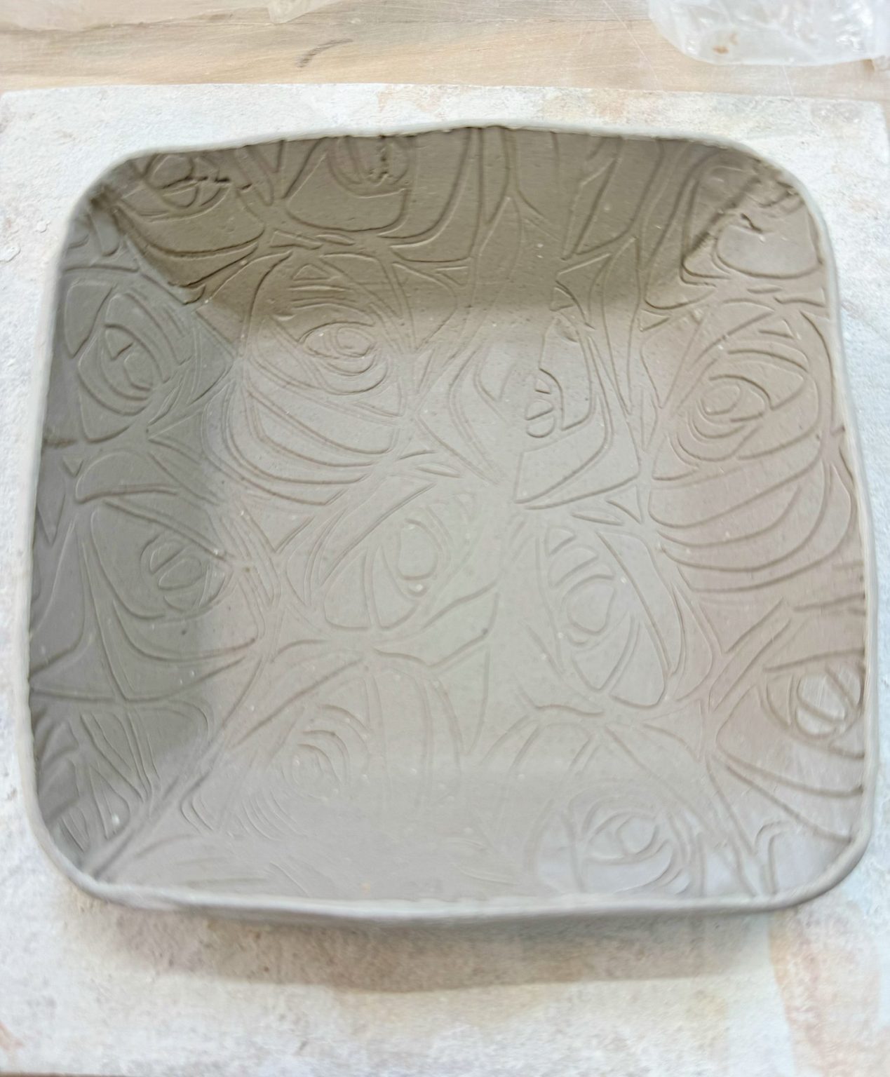 A square, unfired clay dish with a raised abstract swirl pattern covering the surface, placed on a light-colored work surface.