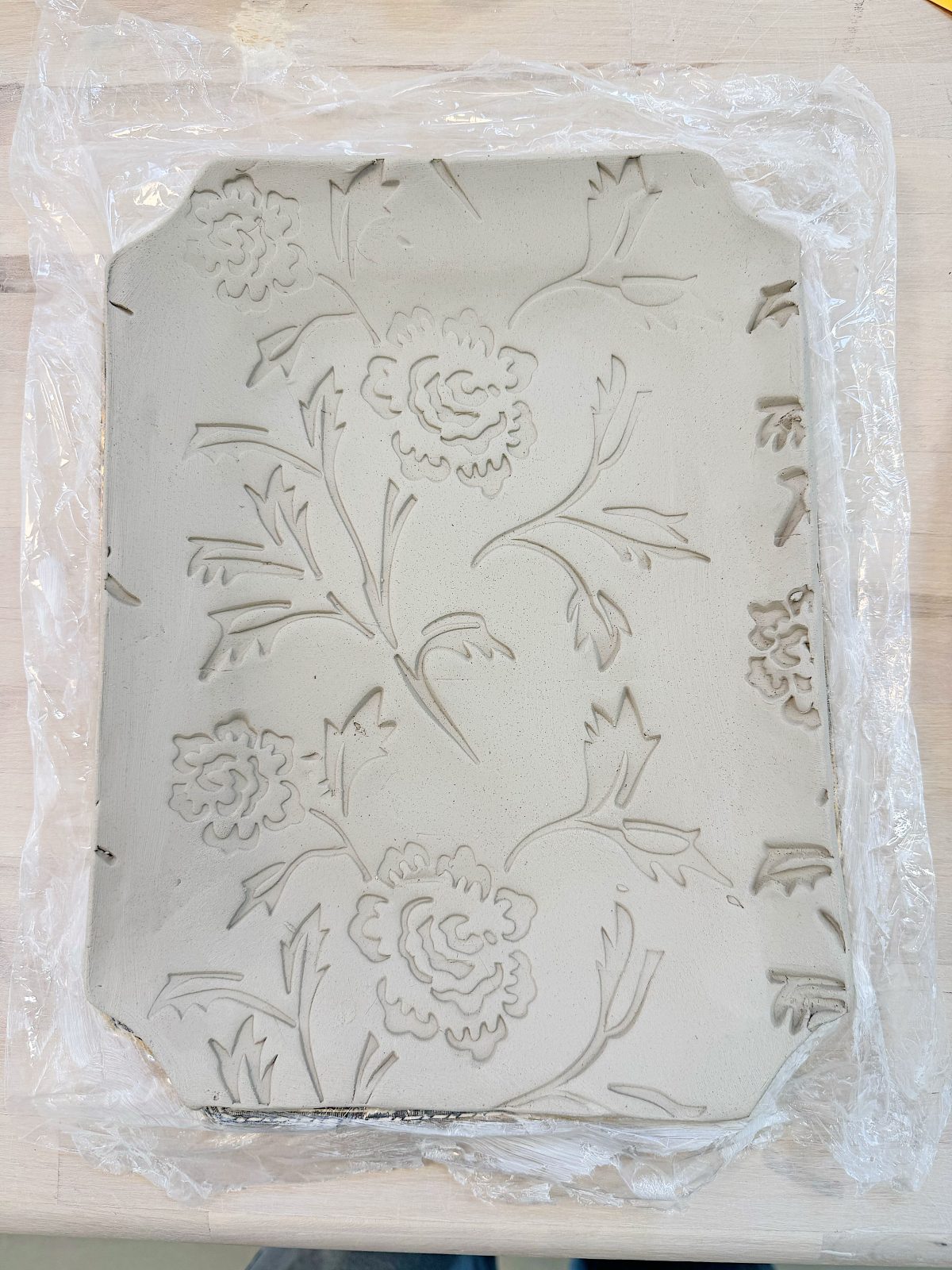 A rectangular clay slab with floral patterns is placed on a wooden surface and partially covered with clear plastic wrap.