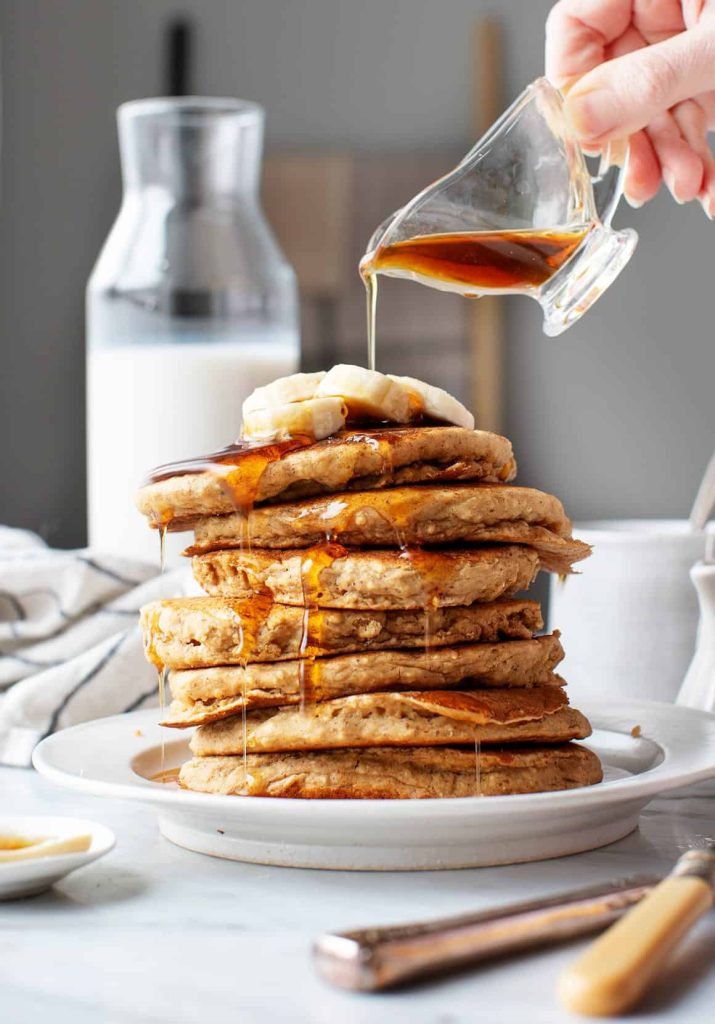 A hand pours syrup over a stack of pancakes topped with banana slices. A carafe of milk is in the background on a marble countertop.