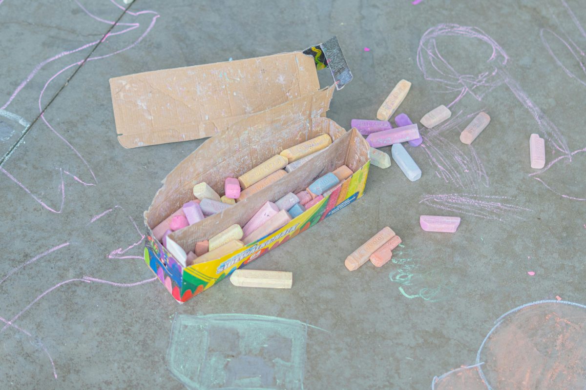 An open box of colored sidewalk chalk lies on pavement surrounded by scattered chalk pieces and childlike chalk drawings.