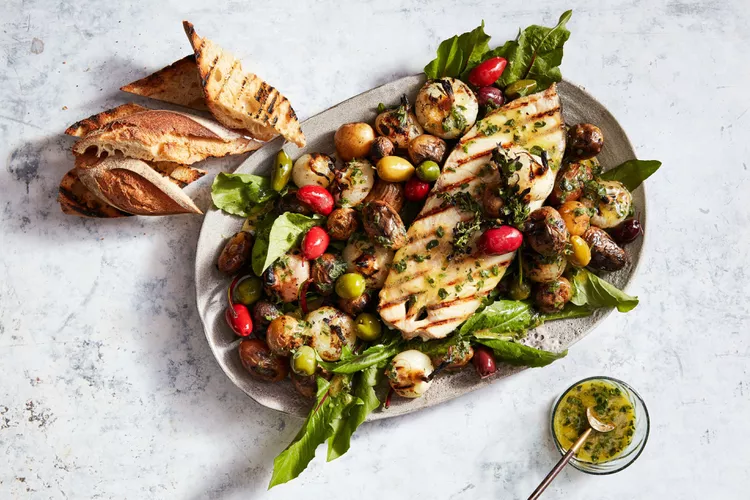 A platter with grilled fish, roasted baby potatoes, olives, cherry tomatoes, leafy greens, grilled bread slices, and a small bowl of herb sauce on the side.