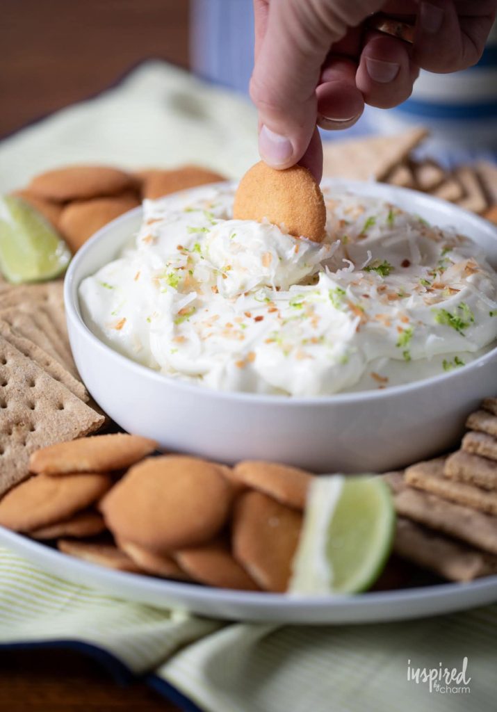A hand dips a vanilla wafer into a bowl of creamy dip, garnished with lime zest and coconut, surrounded by crackers, wafers, and lime wedges.