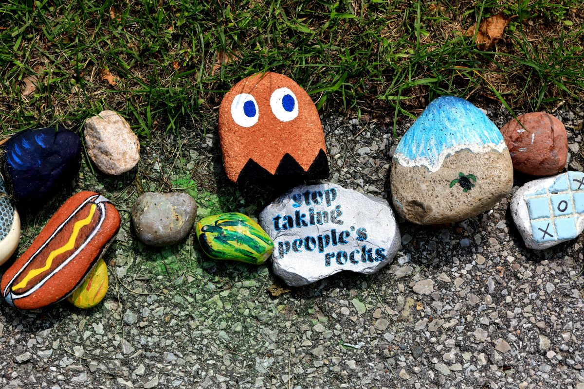 Painted rocks on grass and pavement, including a Pac-Man ghost, a hot dog, a mountain, and one with the text "stop taking people's rocks.