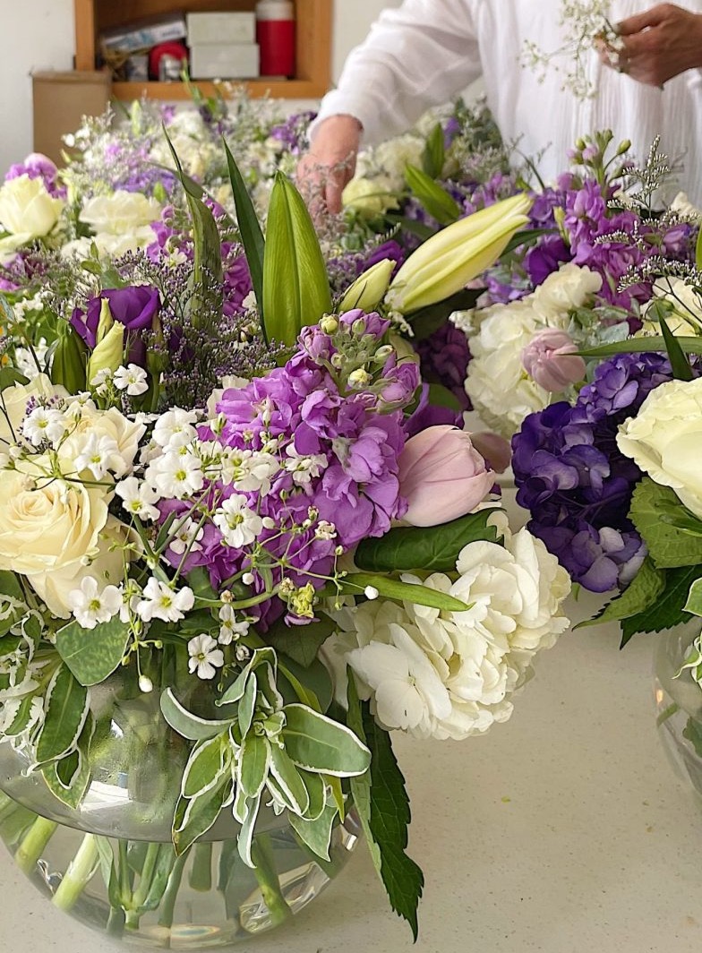 A person arranges assorted fresh summer wedding flowers, including white roses, purple lisianthus, and greenery, in clear glass vases on a table.