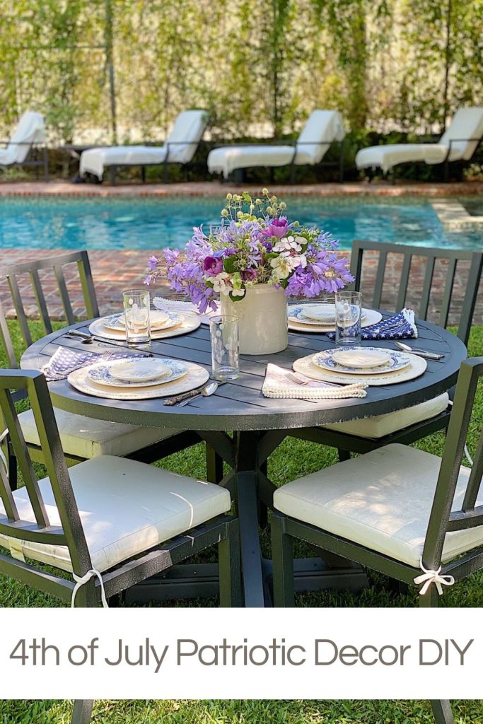 A round outdoor table set for four with plates, glasses, and a vase of purple flowers, near a swimming pool with lounge chairs in the background.