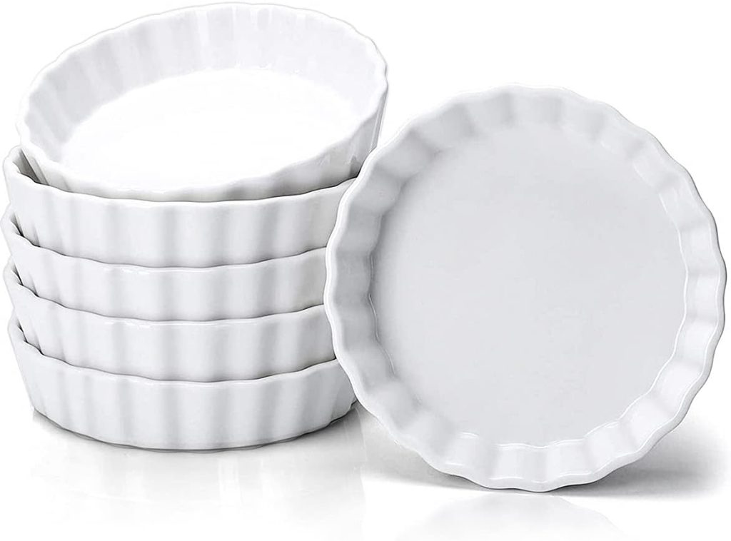 Six white, round, fluted-edge ceramic tart pans are stacked, with one pan leaning against the stack, displayed on a white background—perfect for preparing your favorite mini tart recipe.
