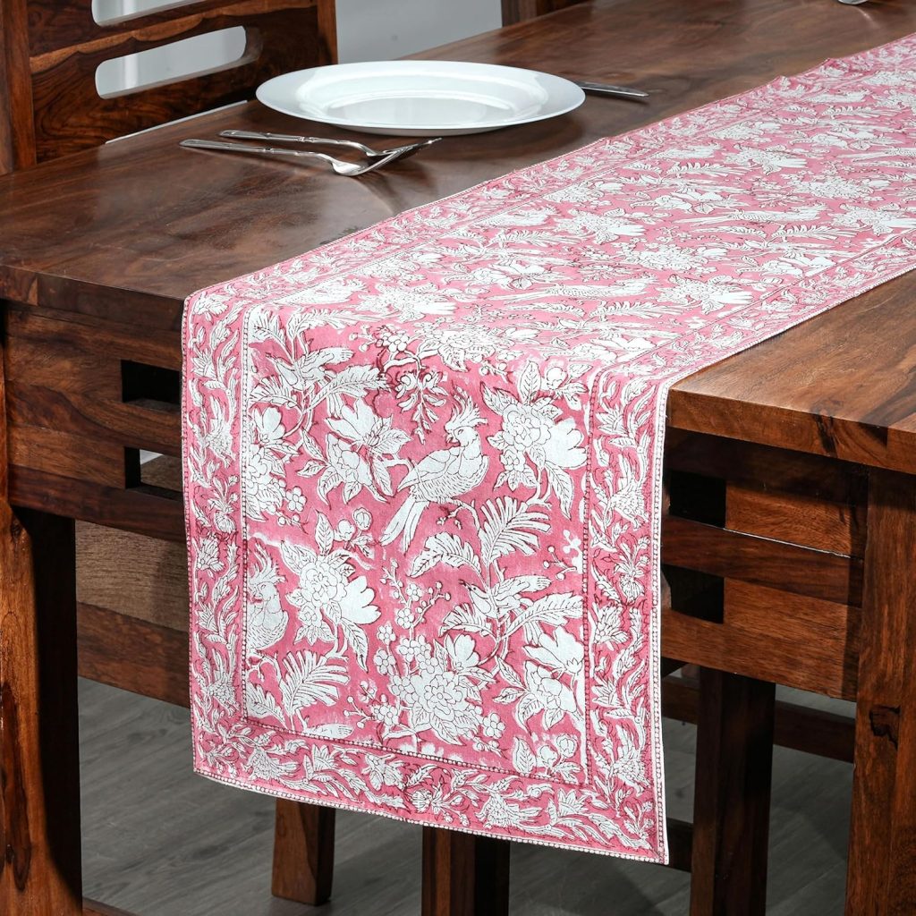 A wooden dining table with a white plate, fork, and knife, decorated with a pink and white floral-patterned table runner—perfect for hosting an elegant dinner party.