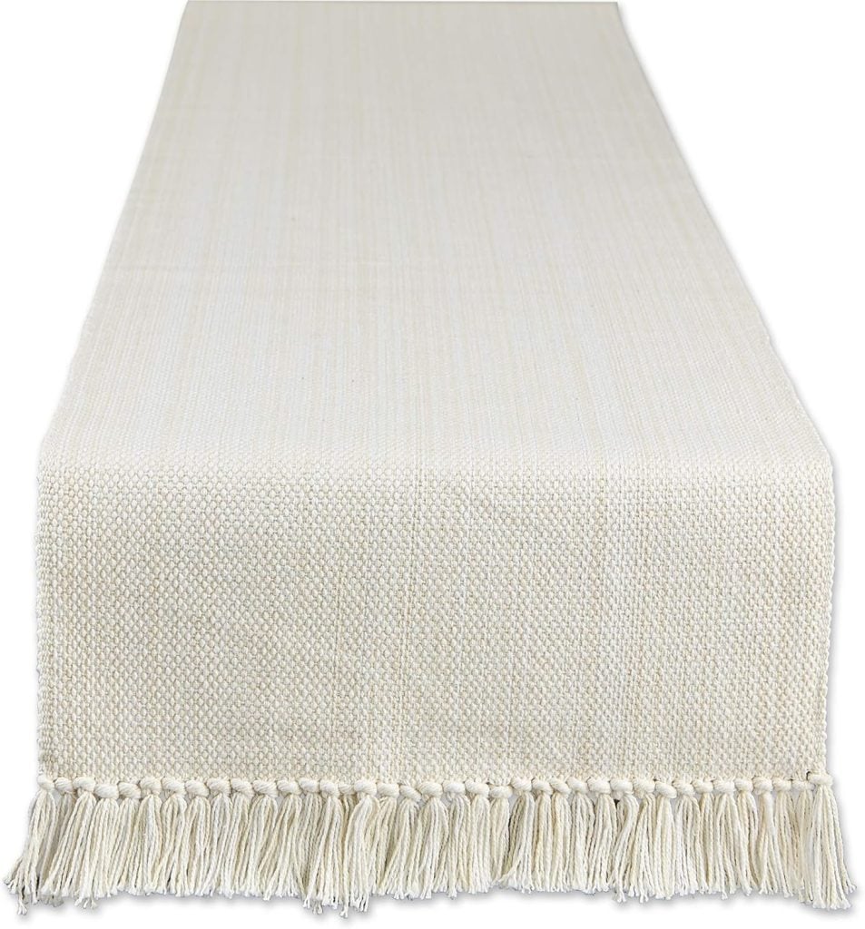 Beige woven table runner with a subtle pattern and fringed edges, ideal for interior designers seeking elegant finishing touches, displayed on a white background.