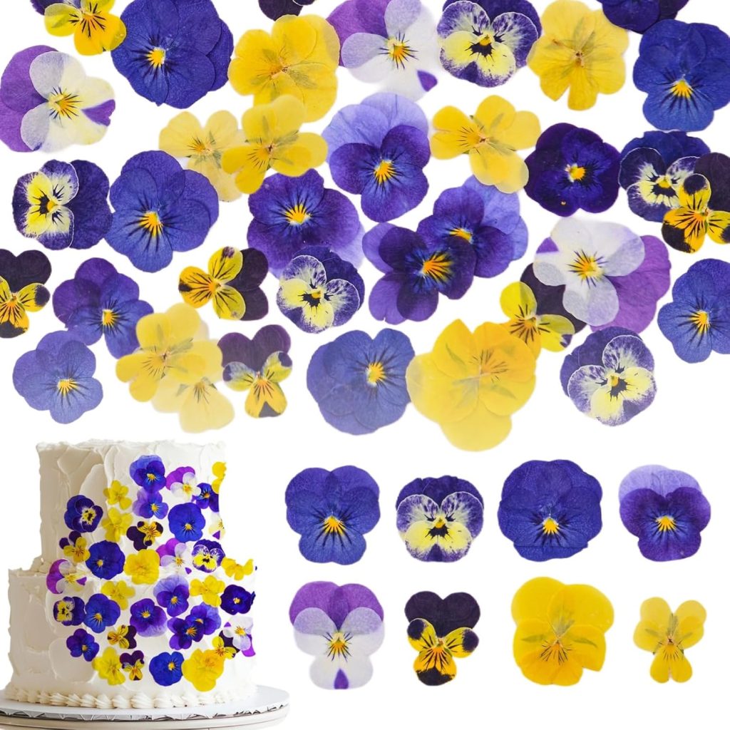 A white cake decorated with purple, yellow, and blue edible pansy flowers sits on a white background, with more pansies and a delicate berry wreath scattered above and around it.