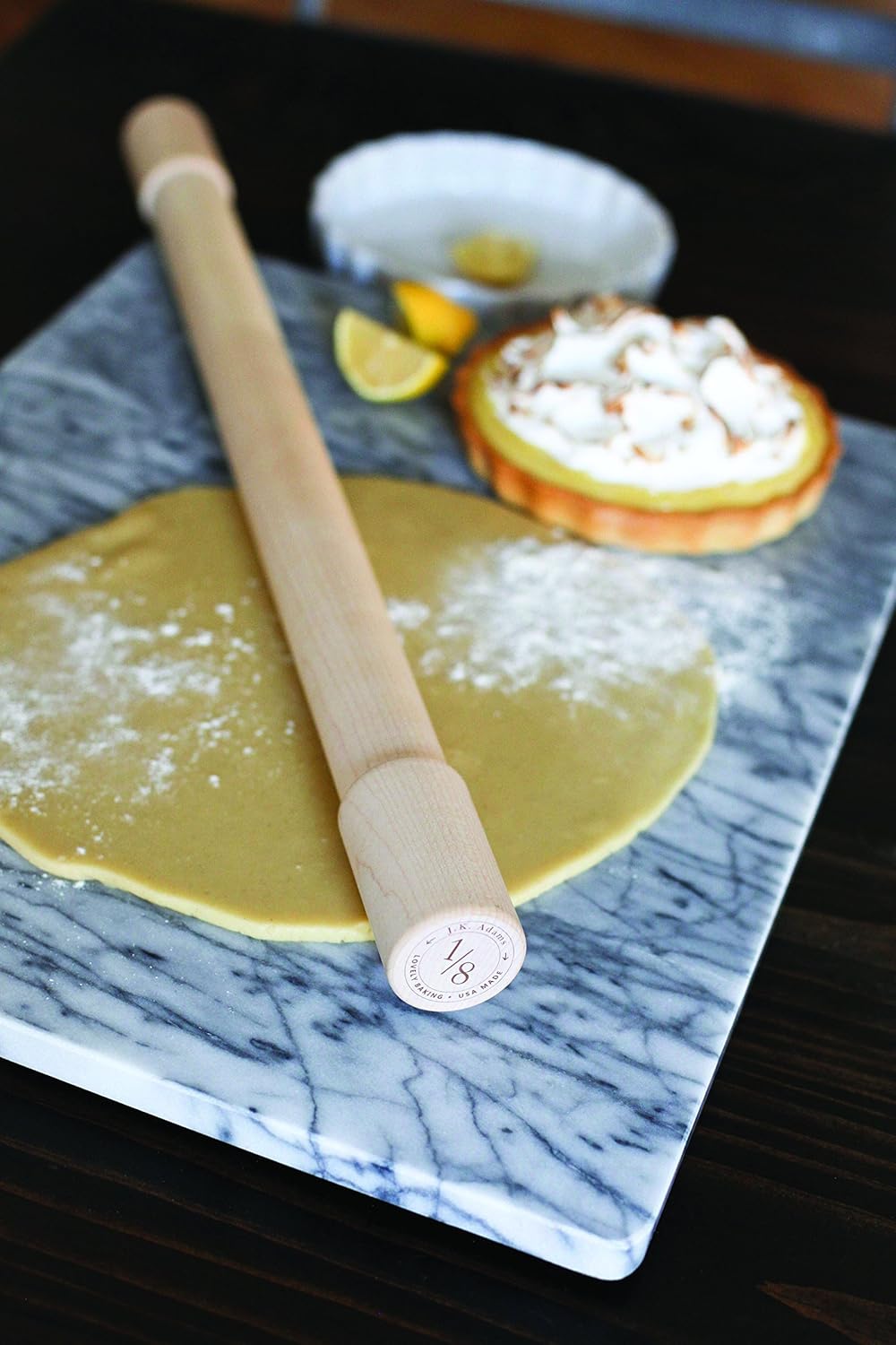 A rolling pin rests on a floured sheet of dough, resembling air dry clay, on a marble board, with a lemon tart and lemon wedges in the background.