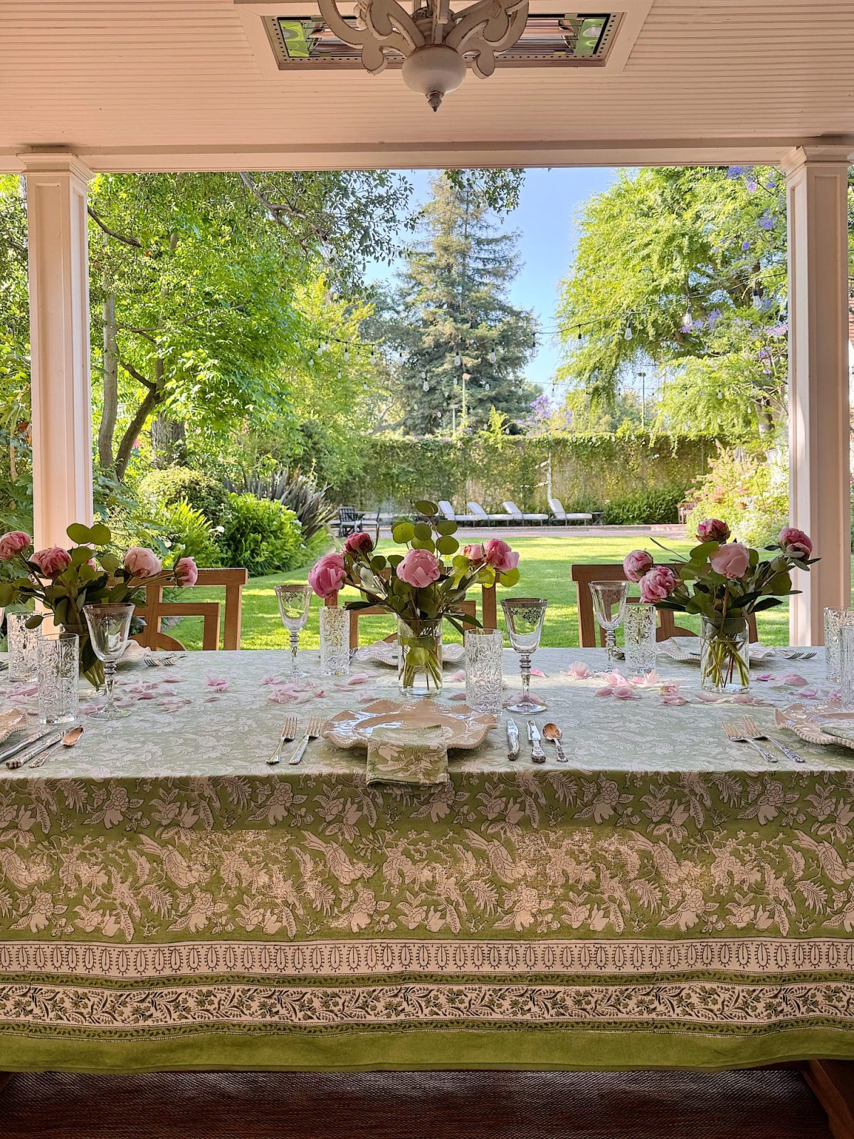 A formal dining table set with floral arrangements, glassware, and silverware is positioned under a porch, overlooking a green garden with trees and lawn chairs in the background.