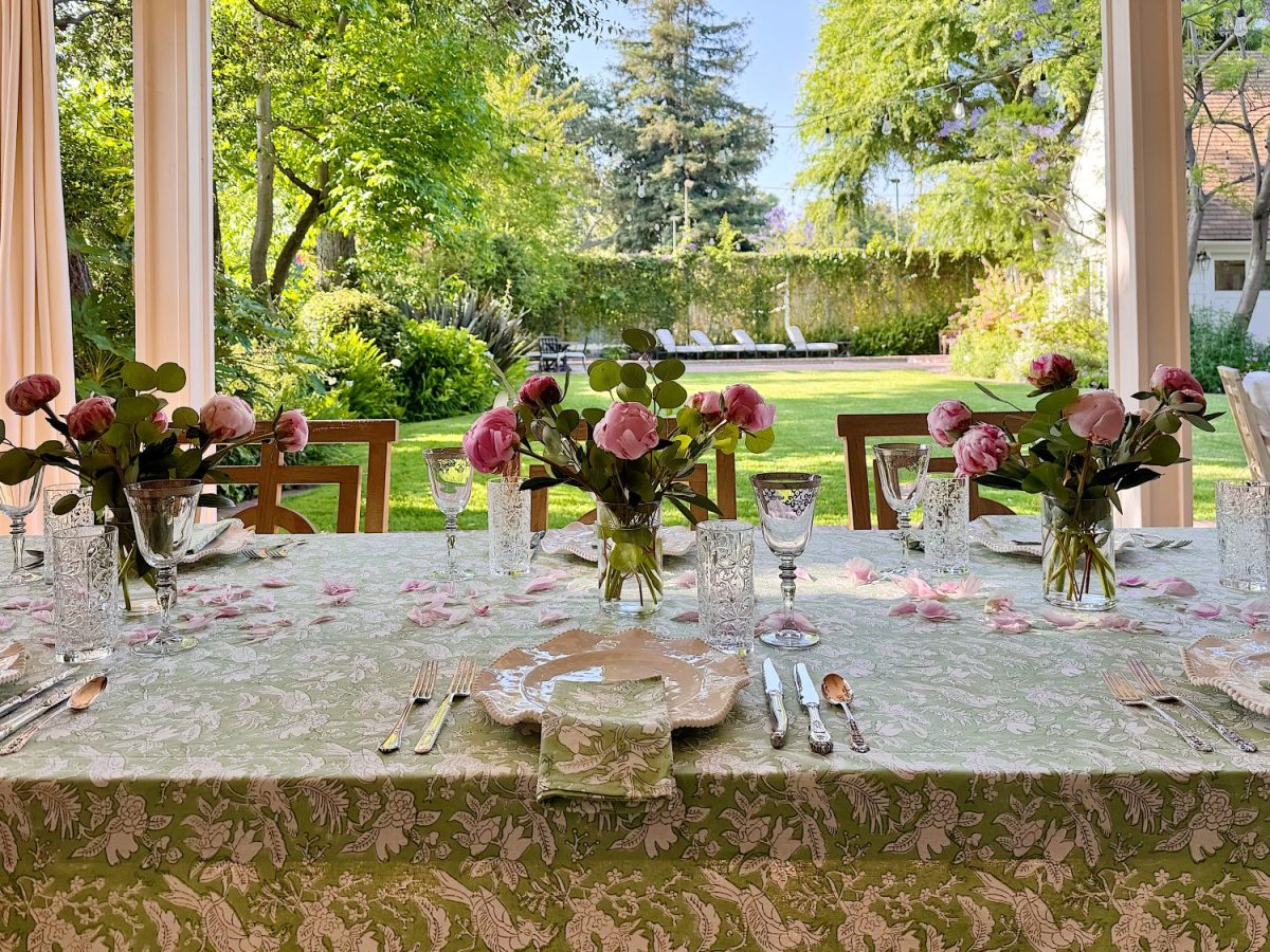 A dining table set with floral-patterned cloth, glassware, cutlery, and vases of pink flowers, positioned by large windows overlooking a green garden.