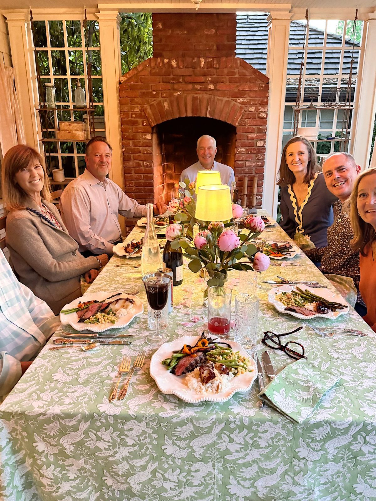 Seven people sit around a dining table set with food and drinks, decorated with flowers and a lamp, in a room with large windows and a brick fireplace.